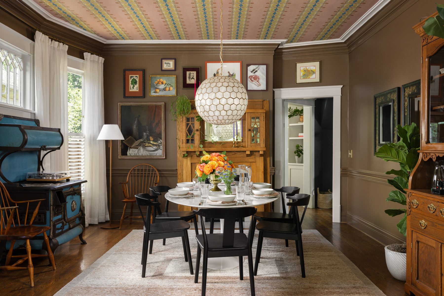 An 1894 dining room with a pink patterned wallpaper on the ceiling, brown walls hung with a collected gallery wall, a capiz honeycomb globe pendant, a marble tulip table with black chairs, an antique blue enamel stove with a turntable, and an oak mantle cabinet