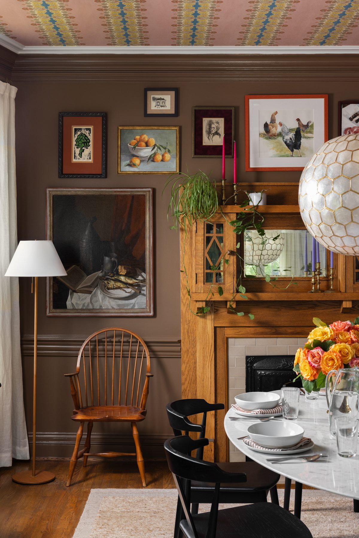 A gallery wall of collected art including an artichoke print, a still life of oranges, and watercolor roosters, above an oak mantle cabinet with a mirror, brass candlesticks, and trailing plants, beside a Windsor chair