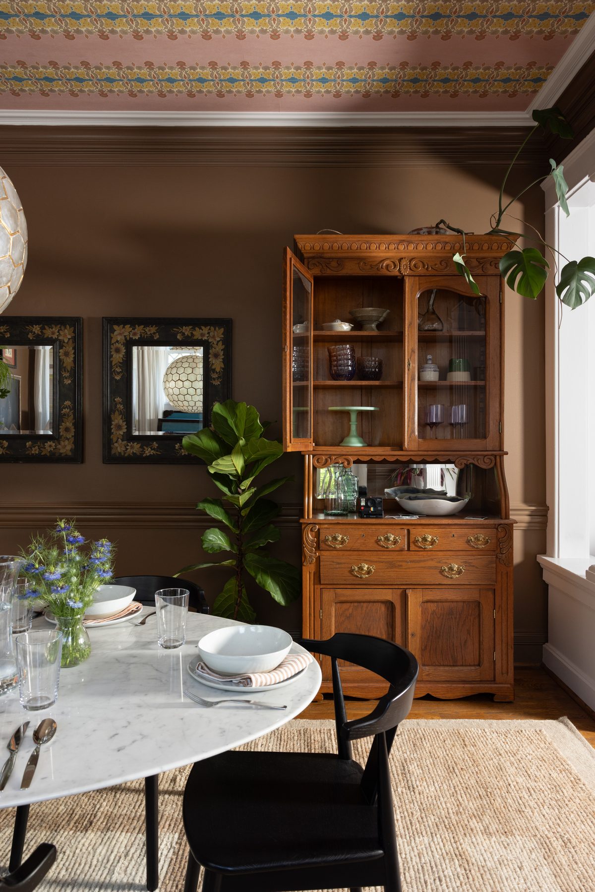 An antique carved oak hutch filled with glassware and a jade cake stand, beside floral-painted mirrors and a fiddle leaf fig, under a pink wallpapered ceiling