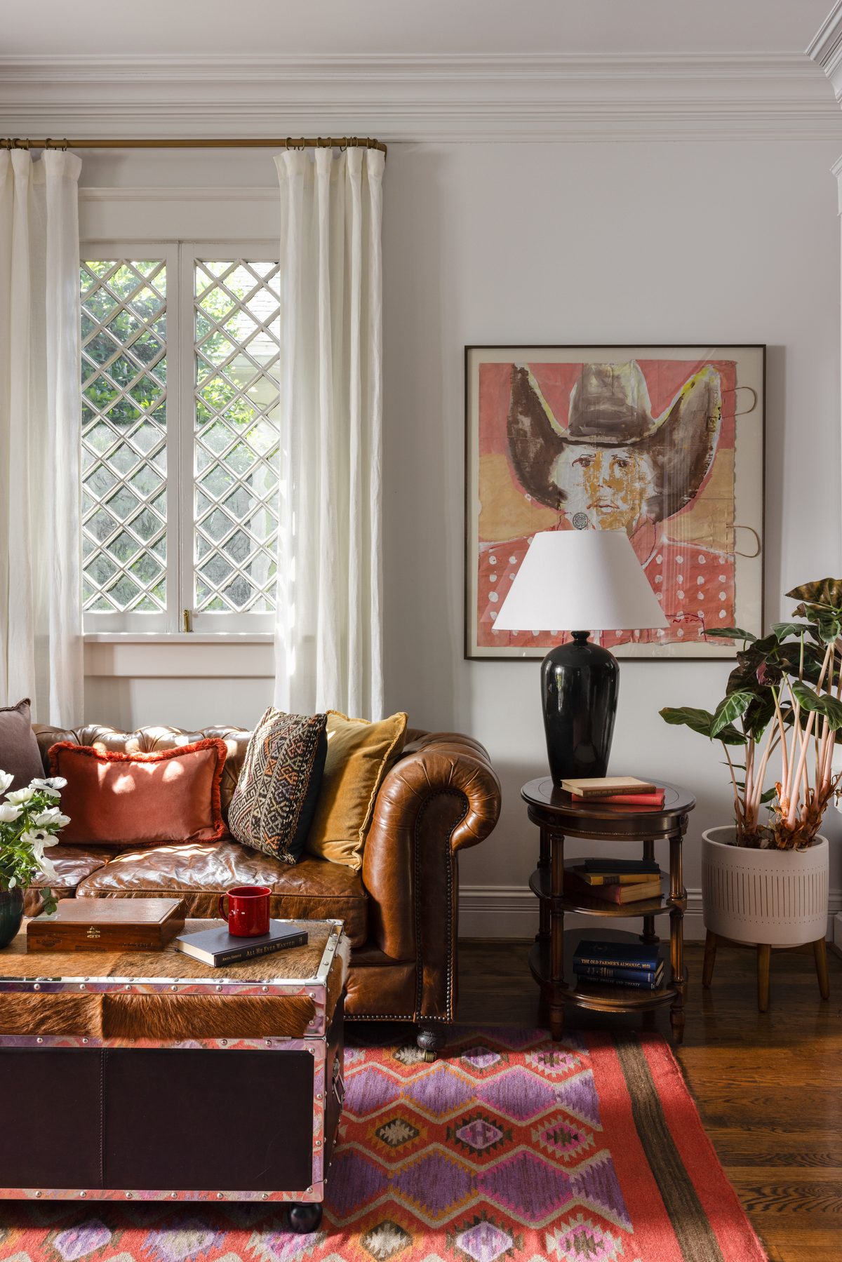 A caramel leather chesterfield sofa with velvet and kilim pillows beneath a large pink cowboy painting, beside a diamond-pane window and a pink kilim rug
