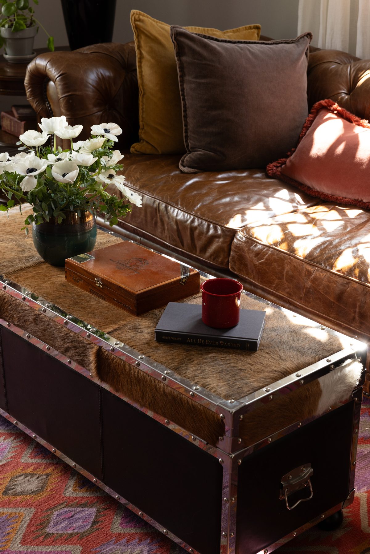 The cowhide trunk coffee table styled with white anemones, a vintage cigar box, and a red enamel mug on a book, in front of the caramel leather chesterfield