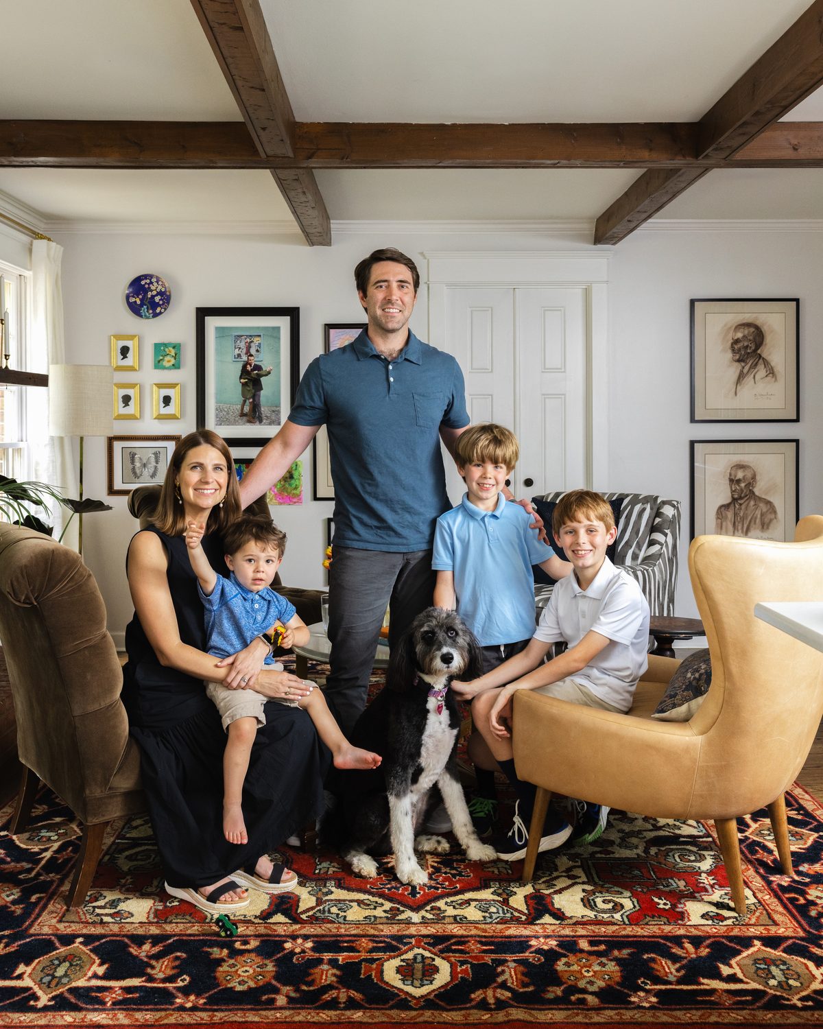 A family of five and their dog in the finished keeping room, surrounded by the four-chair seating arrangement, a gallery wall, and exposed beams