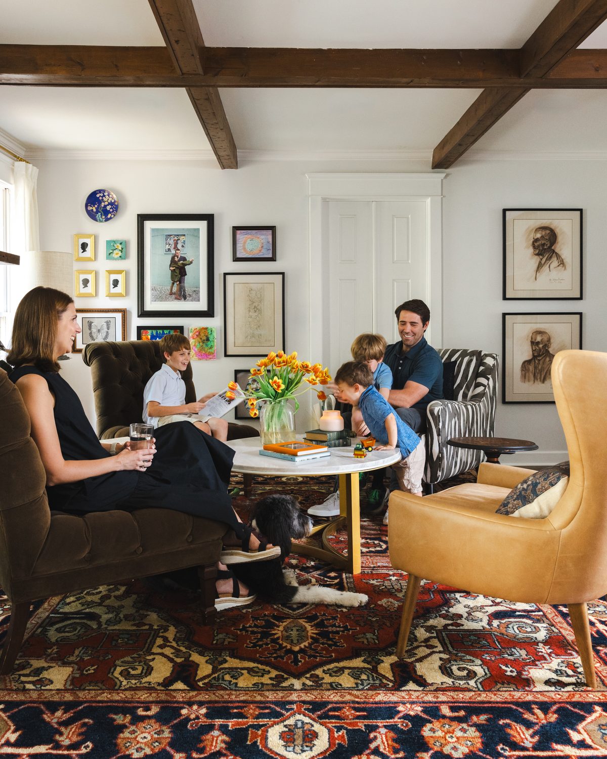 A keeping room with an olive velvet sofa, a grid of six framed vintage prints and textiles, a taupe-painted ceiling, and hanging rugs beside an oak cabinet