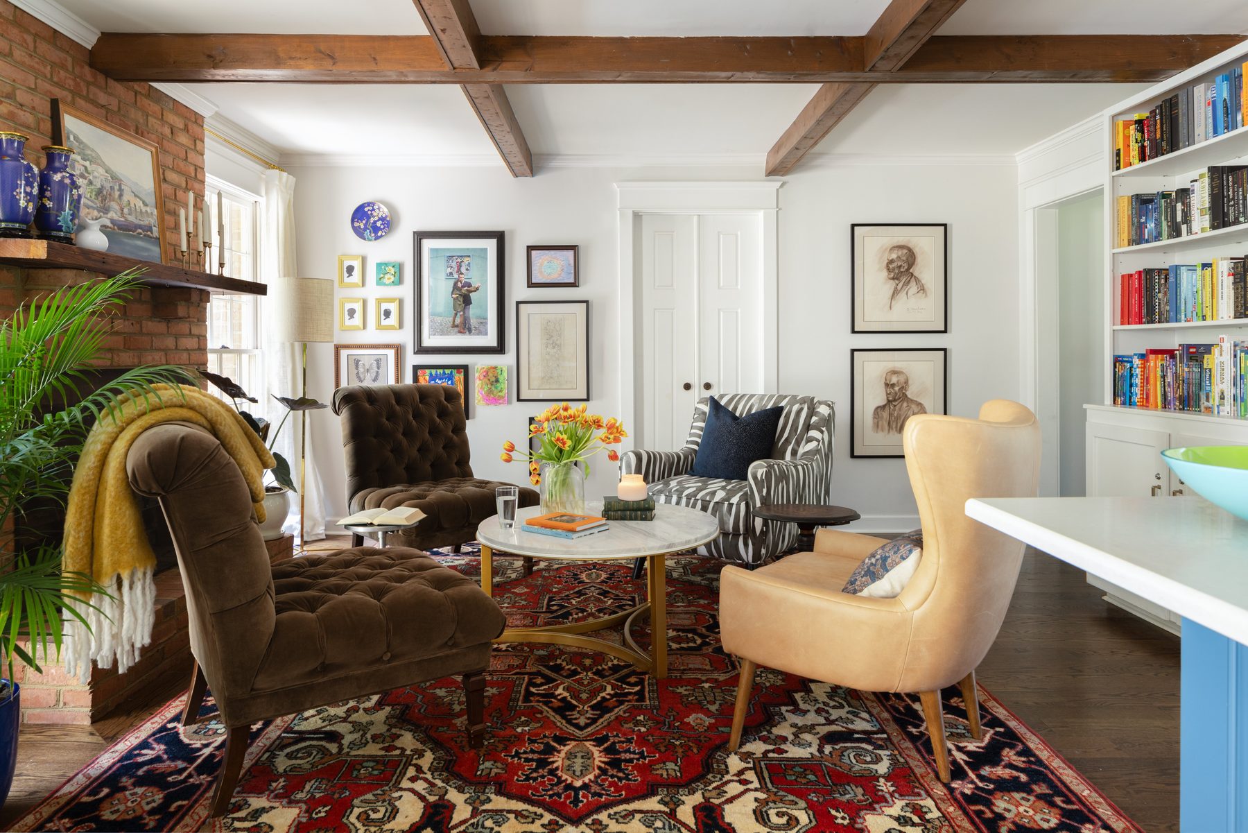 The keeping room off the kitchen: four mismatched chairs around a round marble coffee table on a red Persian rug, with exposed beams, a brick fireplace, and color-arranged bookshelves