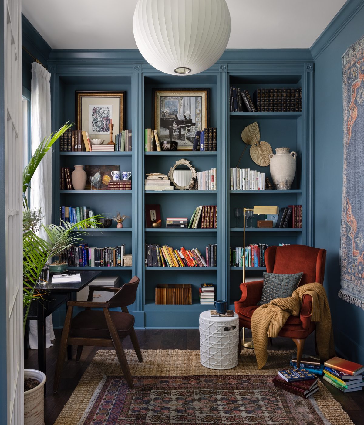 A blue built-in library with a rust velvet wingback chair, layered vintage rugs, and a paper globe pendant, designed by Kristin Keyes Interiors