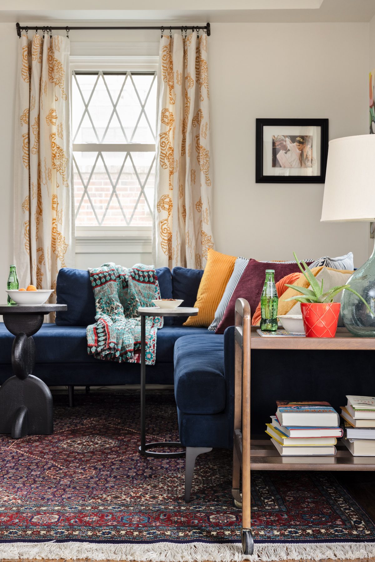 The wood bar cart stacked with books beside the navy sectional, the couple&rsquo;s wedding photo framed above