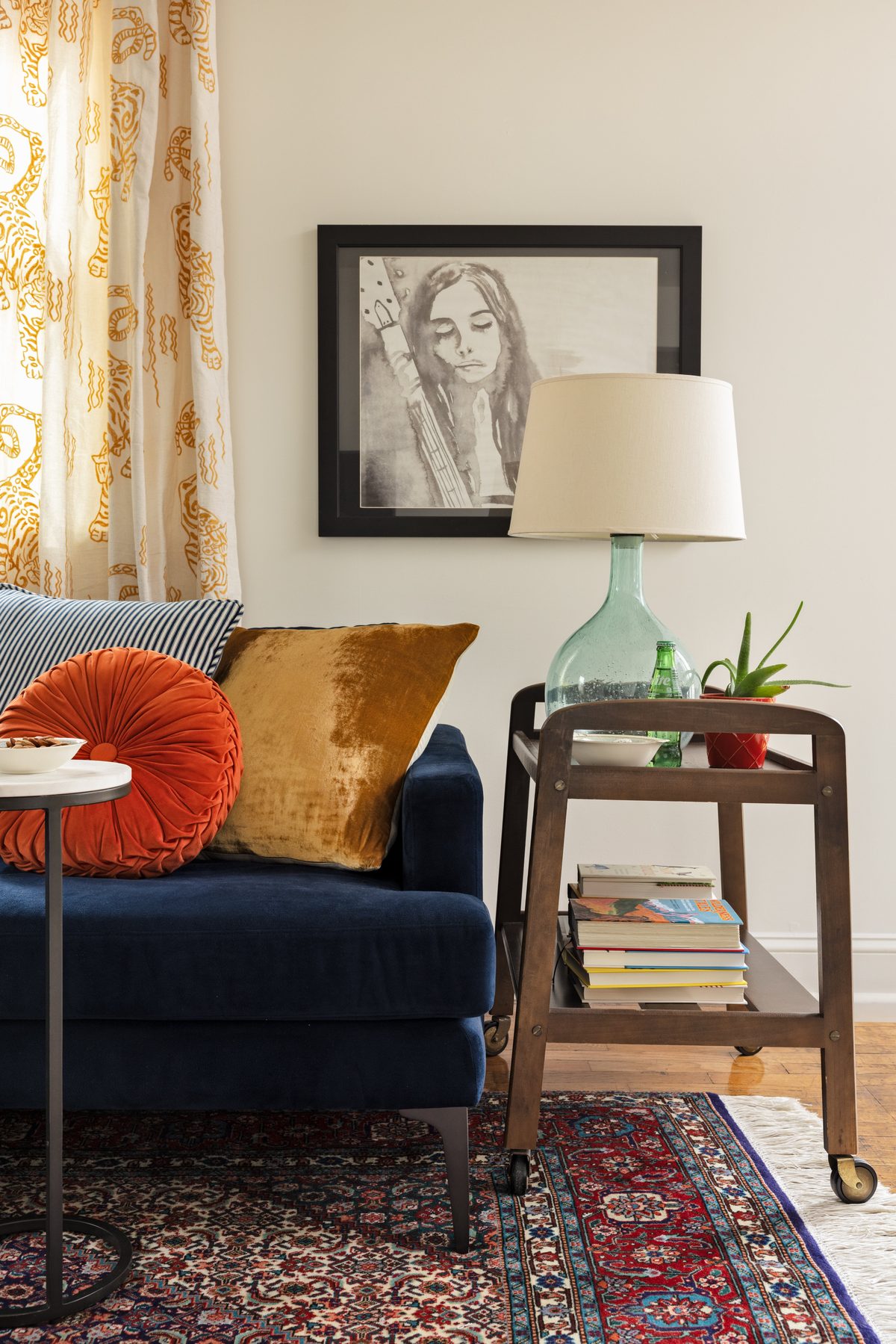 An ink-wash portrait in a black frame above the wood bar cart with a seeded-glass lamp, beside the navy velvet sofa with orange and gold velvet pillows