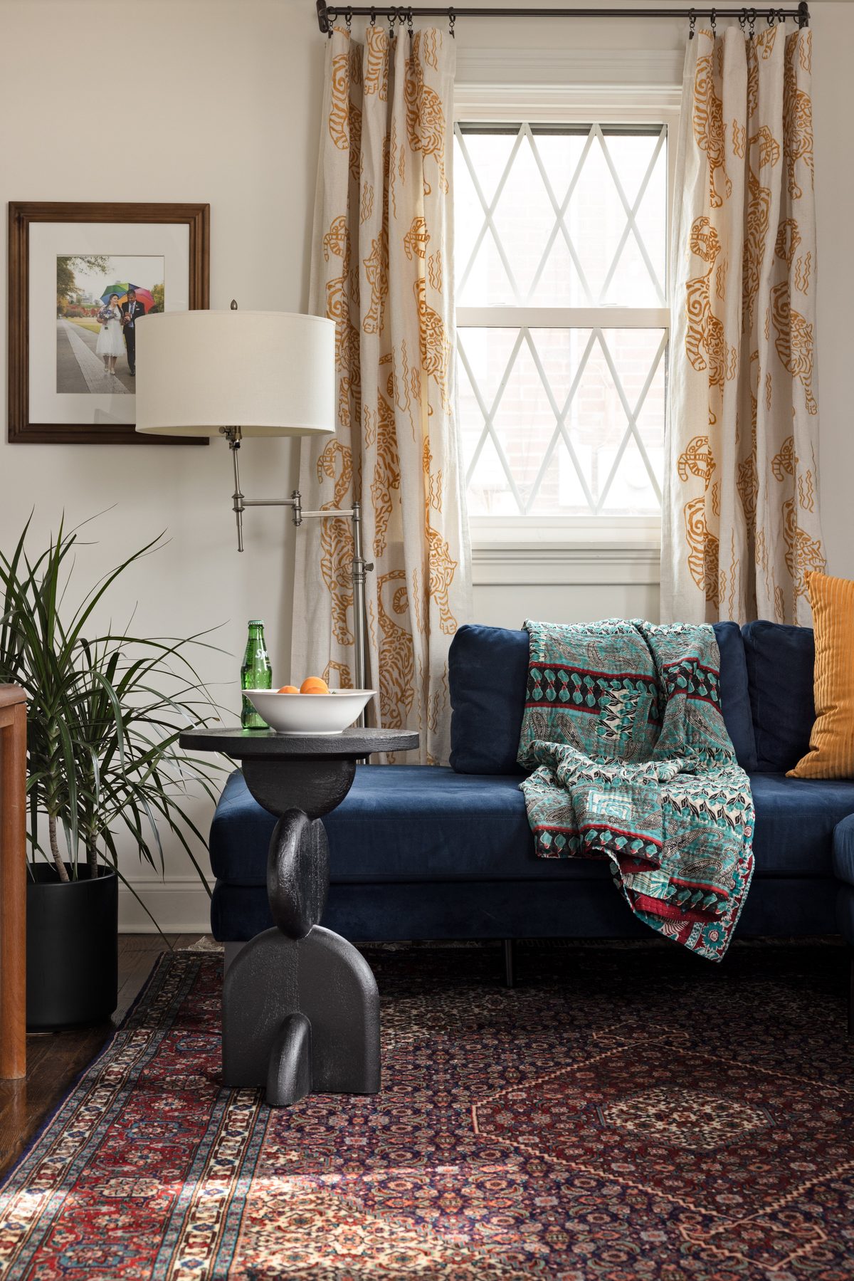 The navy velvet sectional&rsquo;s daybed end with a kantha throw, a black sculptural side table with a bowl of apricots, tiger-print drapery, and the couple&rsquo;s wedding photo on the wall
