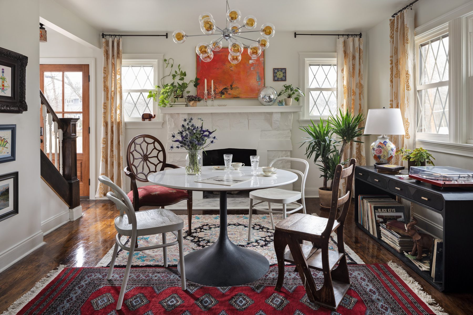 The listening room: a tulip table with mismatched chairs on layered Persian rugs, a whitewashed stone fireplace with an orange abstract painting and a disco ball on the mantle, a sputnik chandelier with amber globes, and a black record console with a turntable