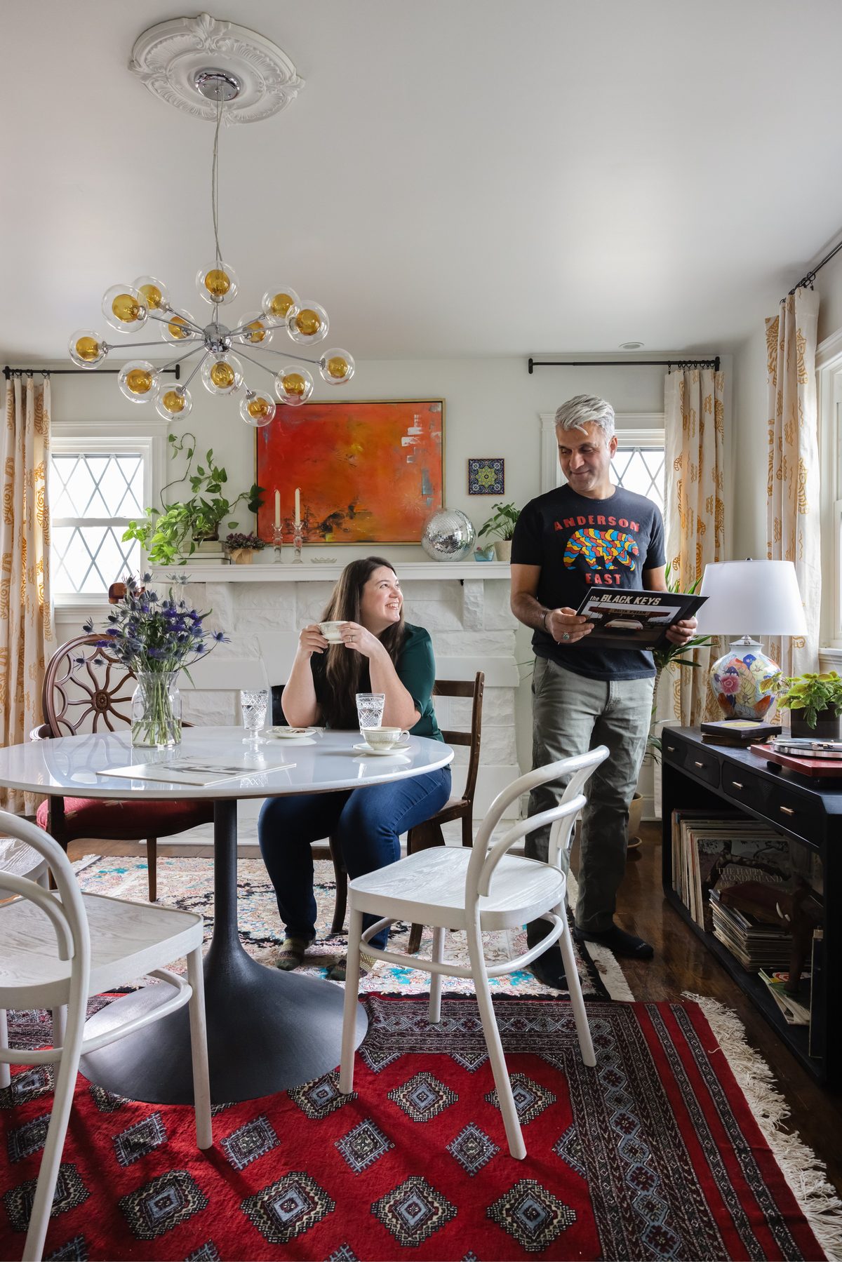 A couple sharing tea and records in a dining room with a tulip table, mismatched chairs, a sputnik chandelier, a whitewashed stone fireplace, and a record console