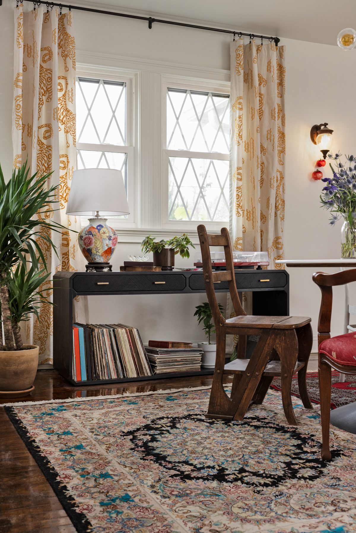 The black record console with a turntable and rows of vinyl below, a tobacco-leaf ginger jar lamp, tiger-print drapery, and an antique library step chair