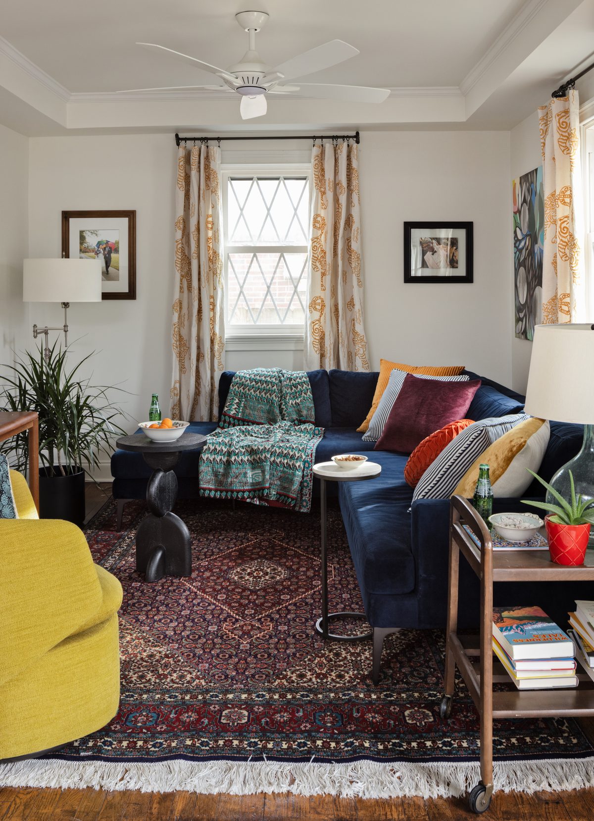The living room with a navy velvet sectional piled with velvet and striped pillows, a kantha throw, a mustard swivel chair, tiger-print drapery, and a layered Persian rug