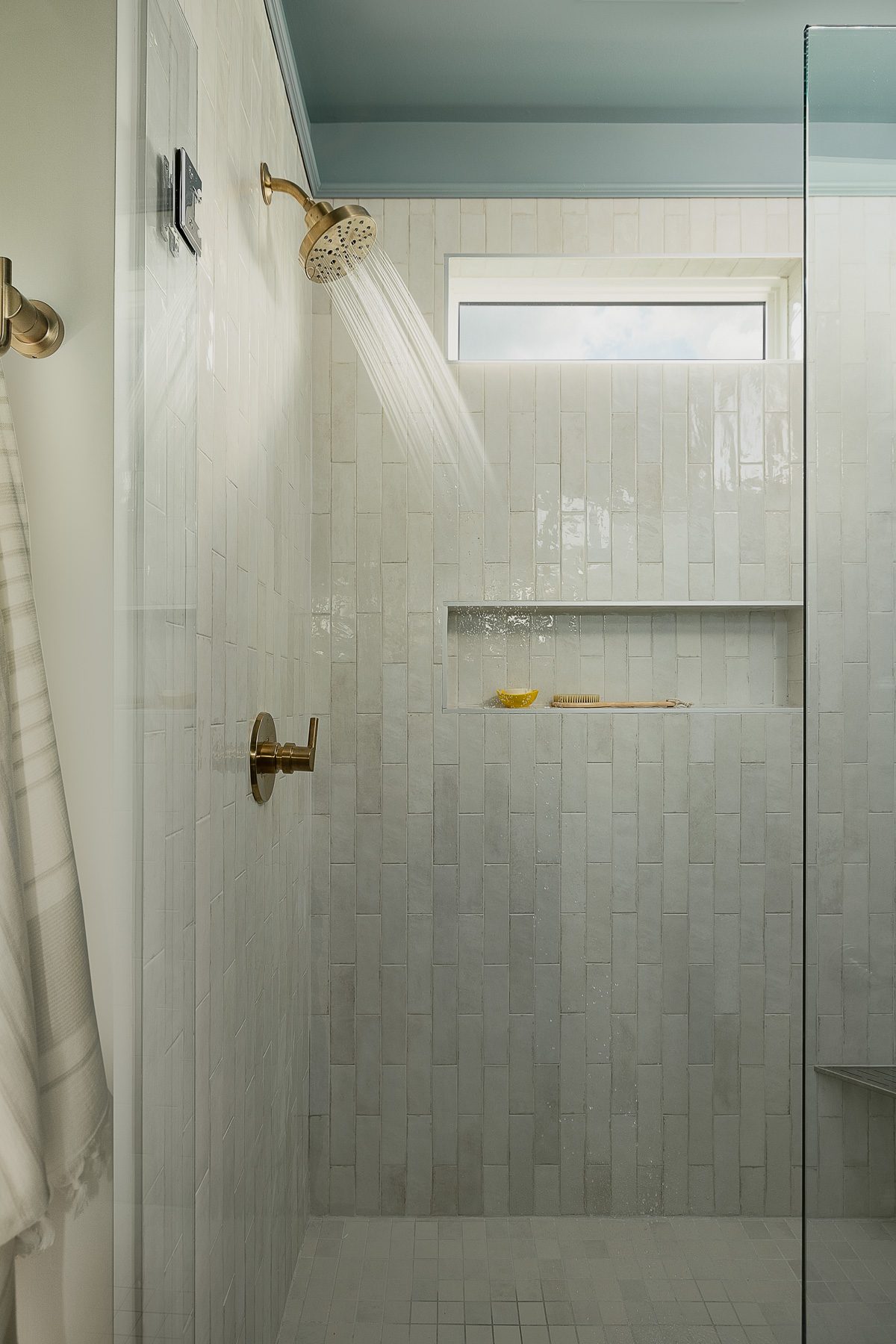A walk-in shower with glossy white stacked tile, brass fixtures, a tiled niche, a transom window, and a soft blue ceiling
