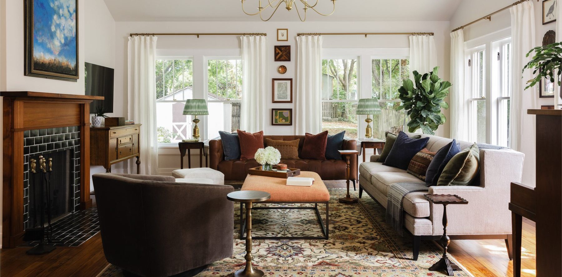 A great room with two facing sofas, an orange linen ottoman, a dark-green-tiled fireplace with a large blue landscape painting, a stack of collected art between two windows, and a brass chandelier