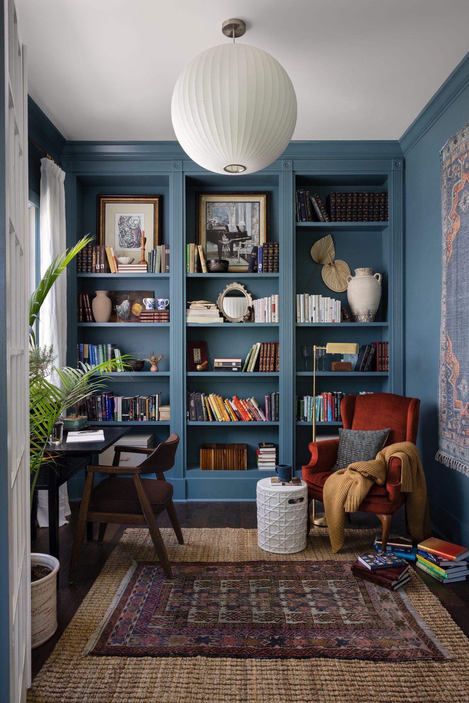 A study with deep blue built-in bookshelves, a rust velvet wingback chair, and a vintage rug hung on the wall