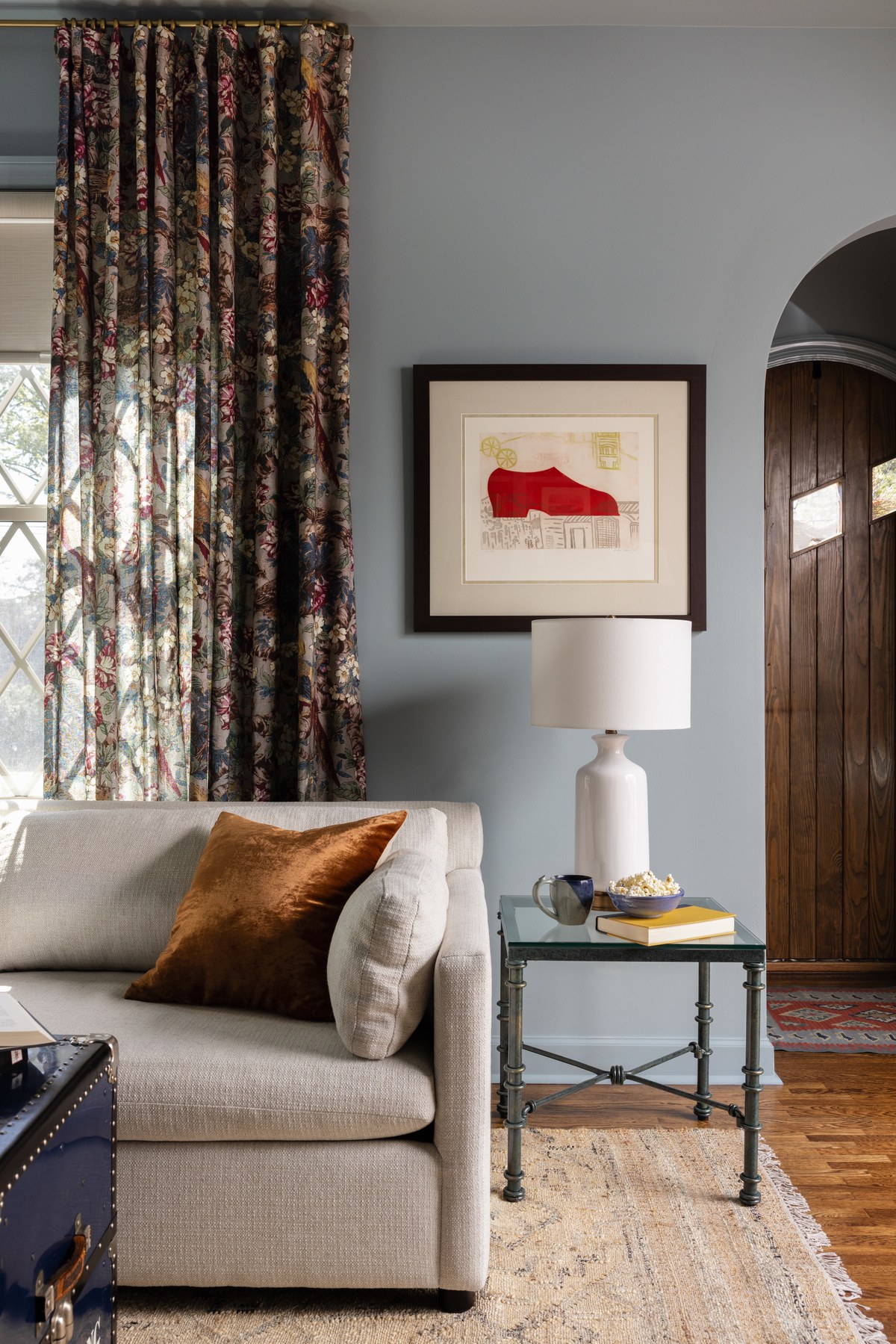 A sunlit corner of the Secret Door sitting room: floral drapery, a red abstract print above a white table lamp, and a rust velvet pillow on the cream sofa