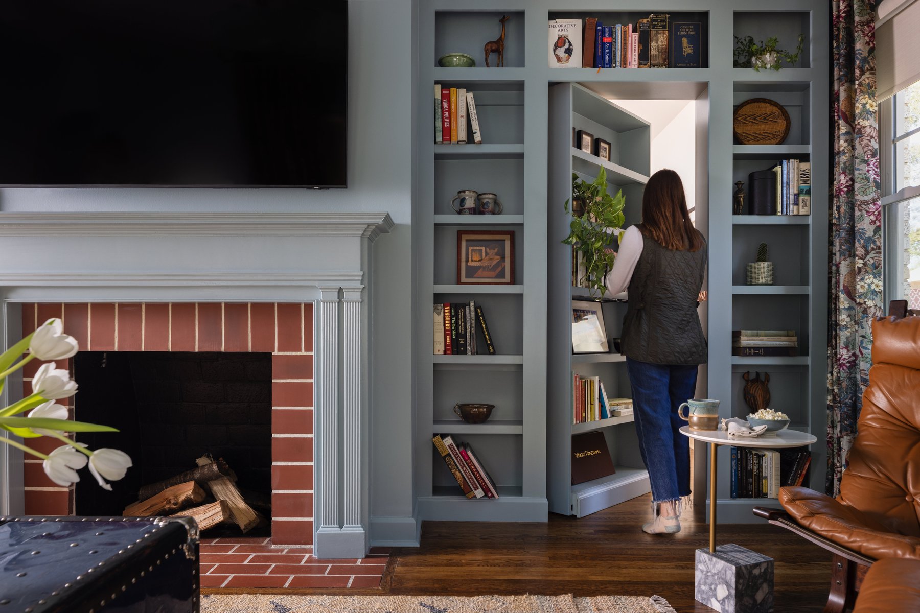 A woman stepping through the open hidden bookcase door in the blue-gray library wall, beside the fireplace