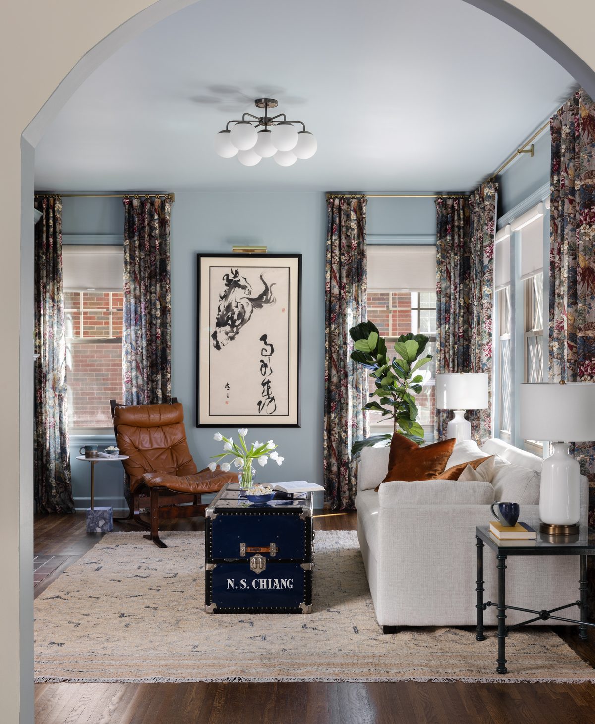 A blue sitting room seen through an arched doorway, with a leather lounge chair, a calligraphy horse artwork, and a vintage trunk coffee table