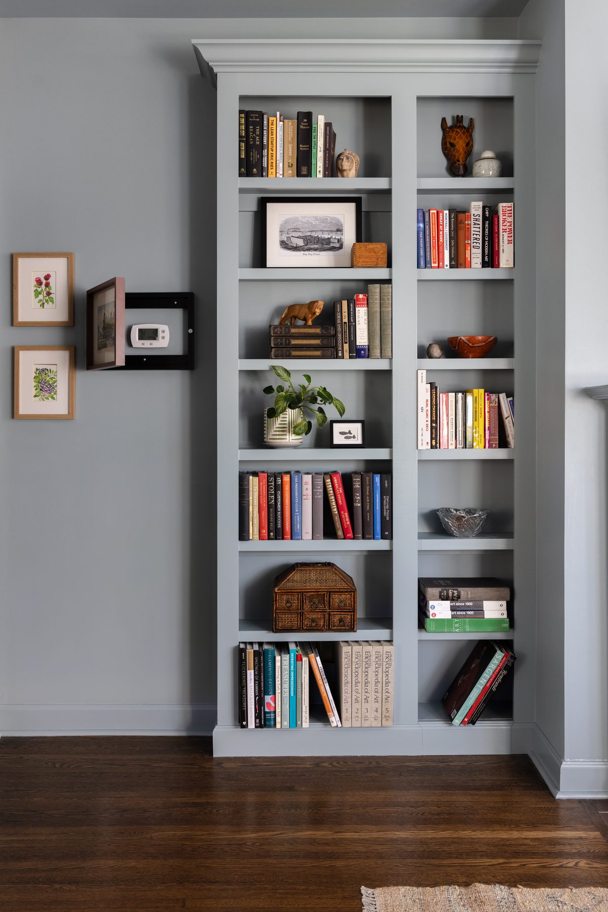 A small hinged picture frame on the wall swung open to reveal the thermostat hidden behind it, beside framed botanical prints