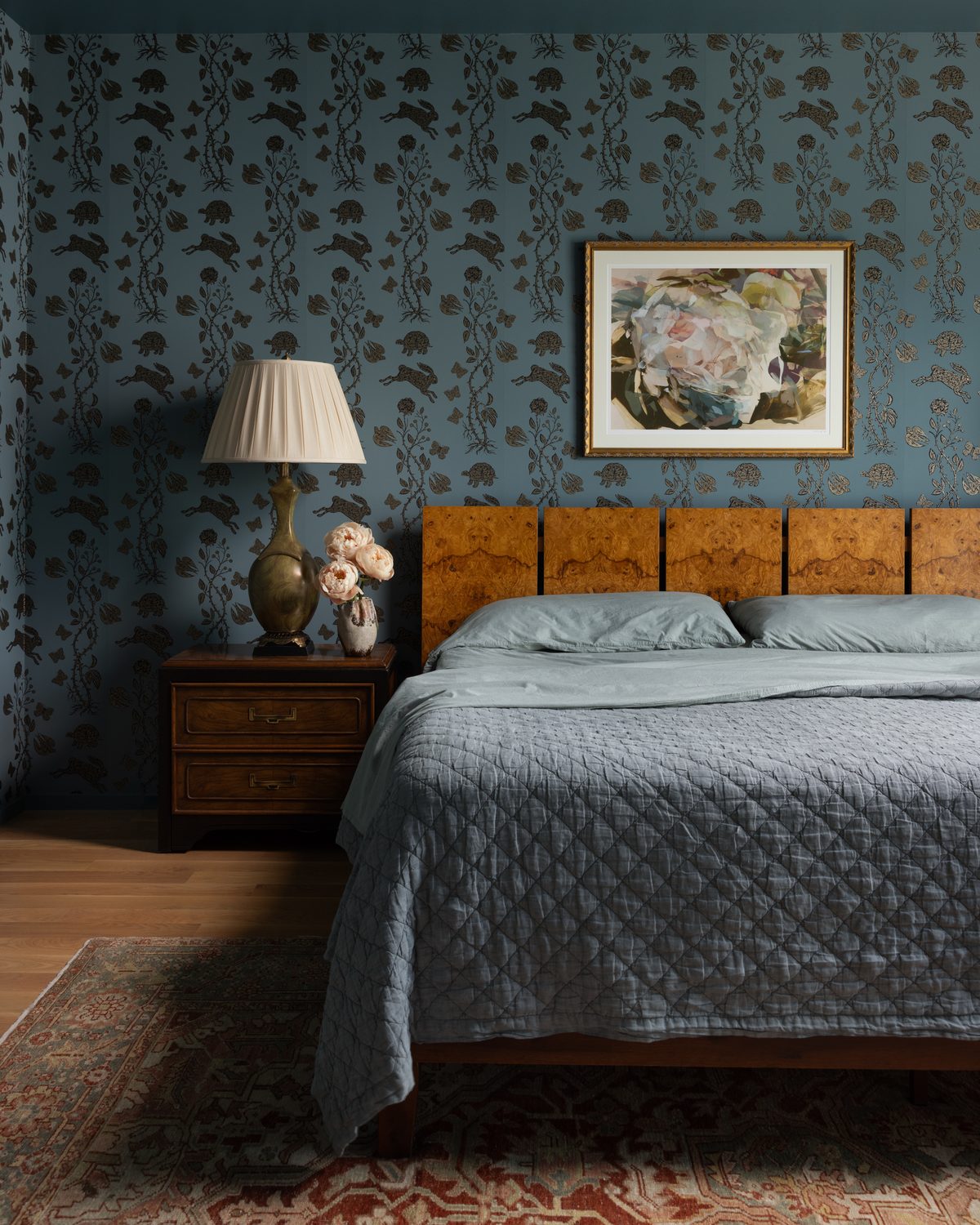 A bedroom with hare-and-turtle wallpaper on slate blue walls, a burl wood headboard above quilted blue bedding, a brass lamp and peonies on a vintage nightstand, a gold-framed abstract painting, and a vintage rug
