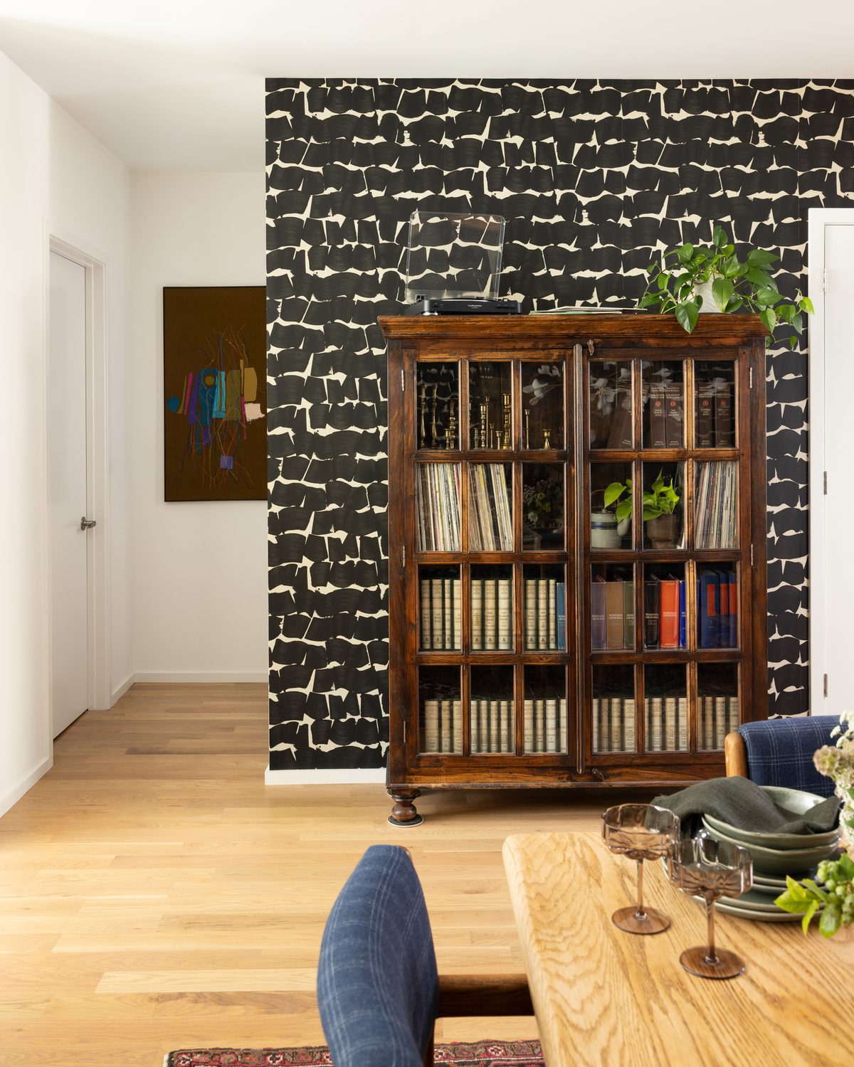 Across the same room: a vintage wood-and-glass cabinet holding records, books, and brass candlesticks against the brushstroke wallpaper, with a turntable on top and the oak dining table in the foreground