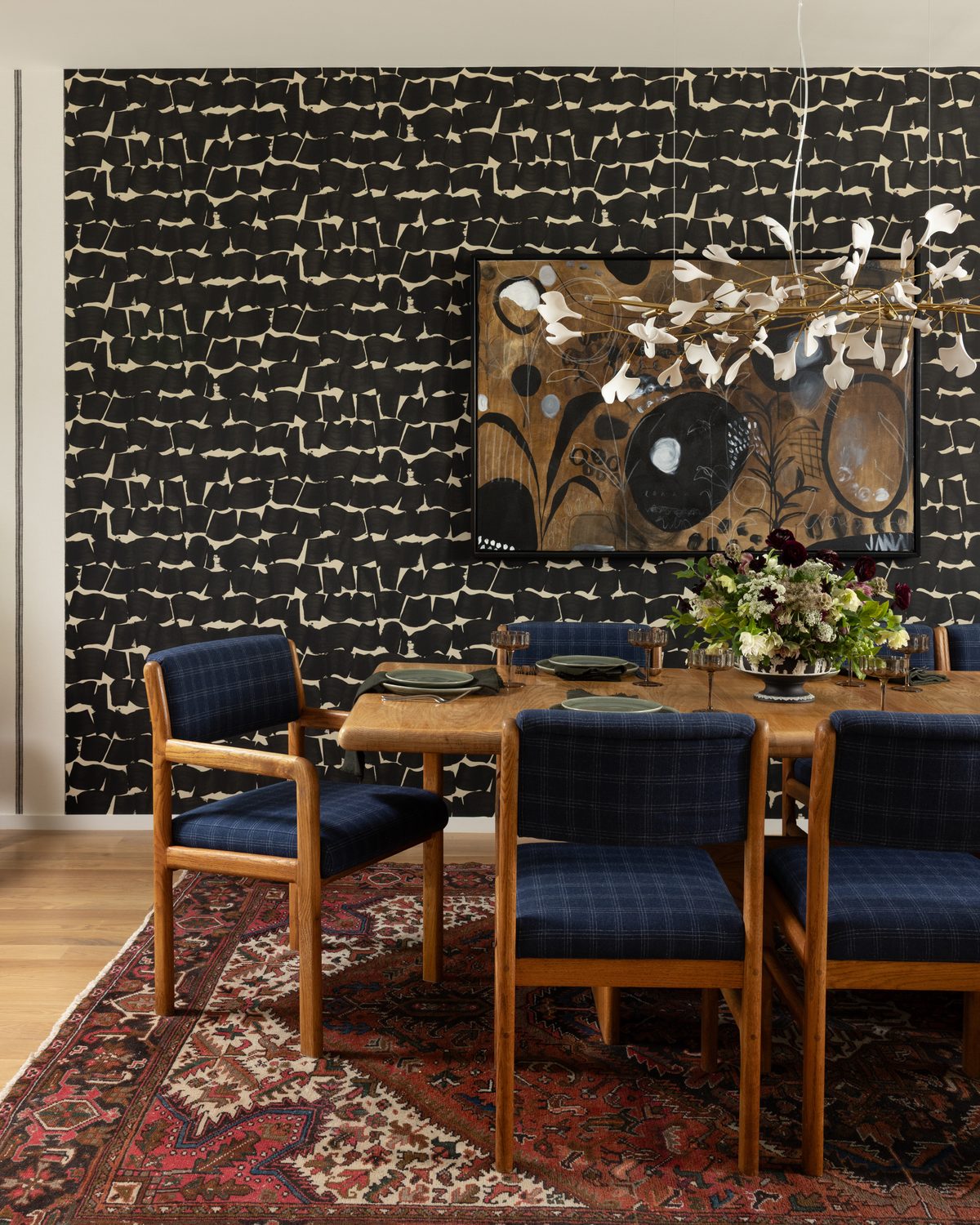 The dining room: black brushstroke wallpaper behind an oak table set with green plates and dark florals, navy plaid chairs, an abstract painting, a porcelain-petal chandelier, and a vintage red rug