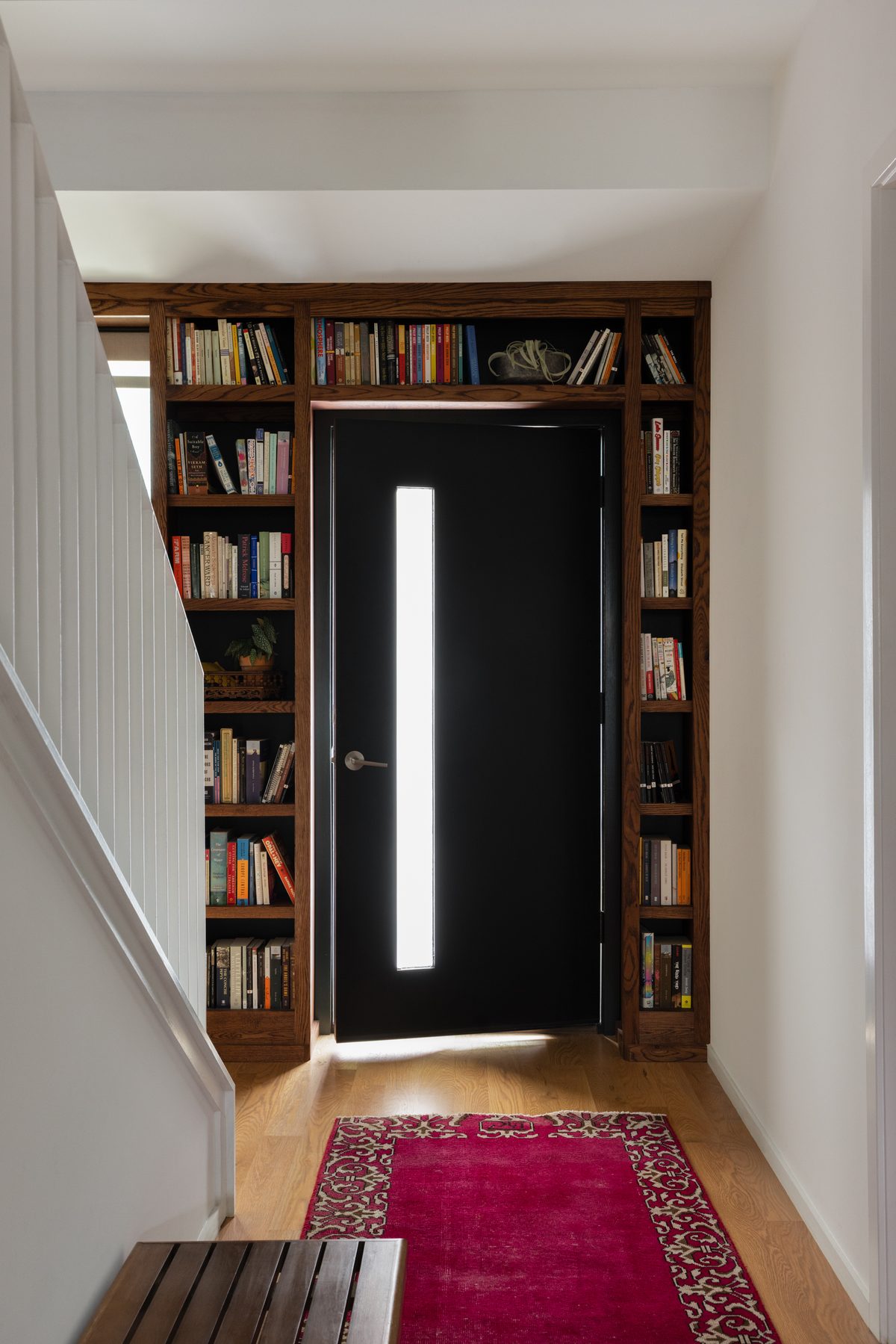 A black front door swung open with light spilling through, framed by floor-to-ceiling bookshelves, with a magenta vintage runner leading to it