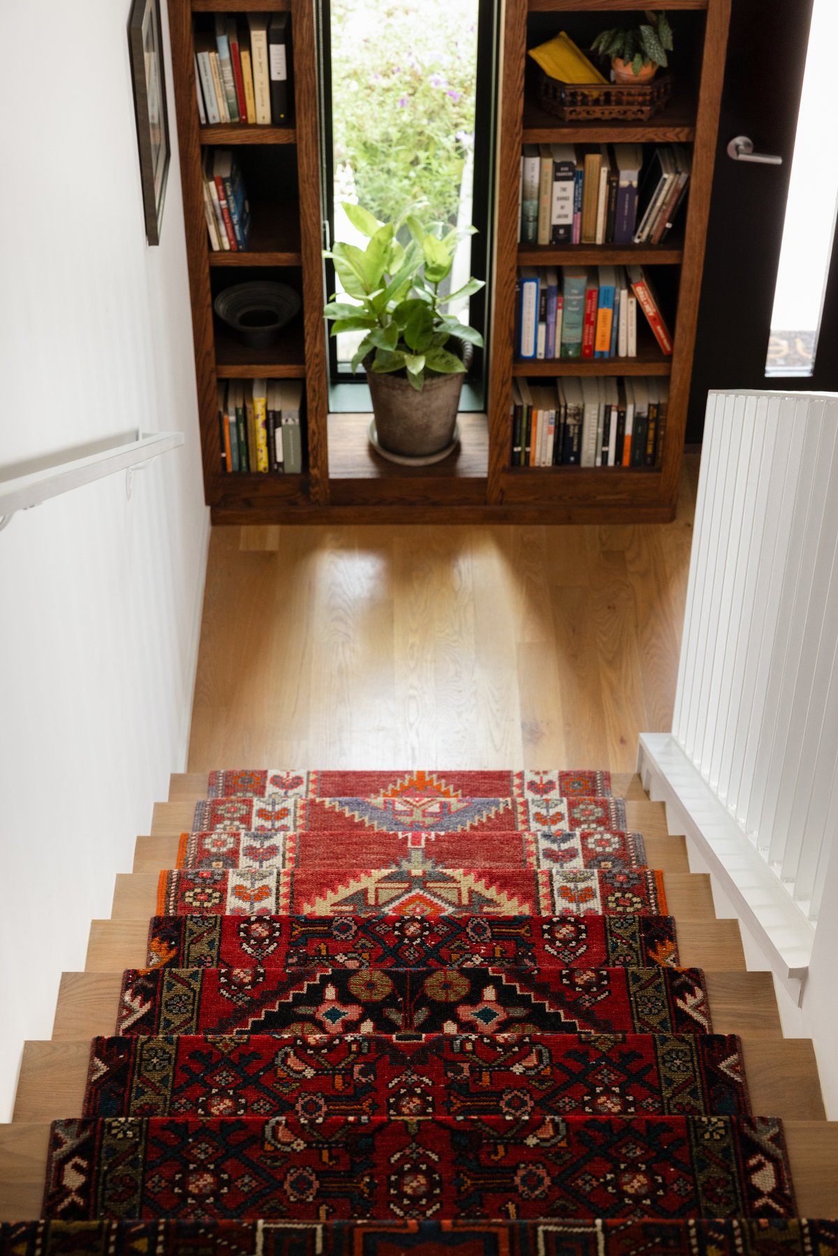 Looking down the rug-runner stairs toward the front door, where built-in walnut bookshelves frame the glass door and a potted plant catches the light