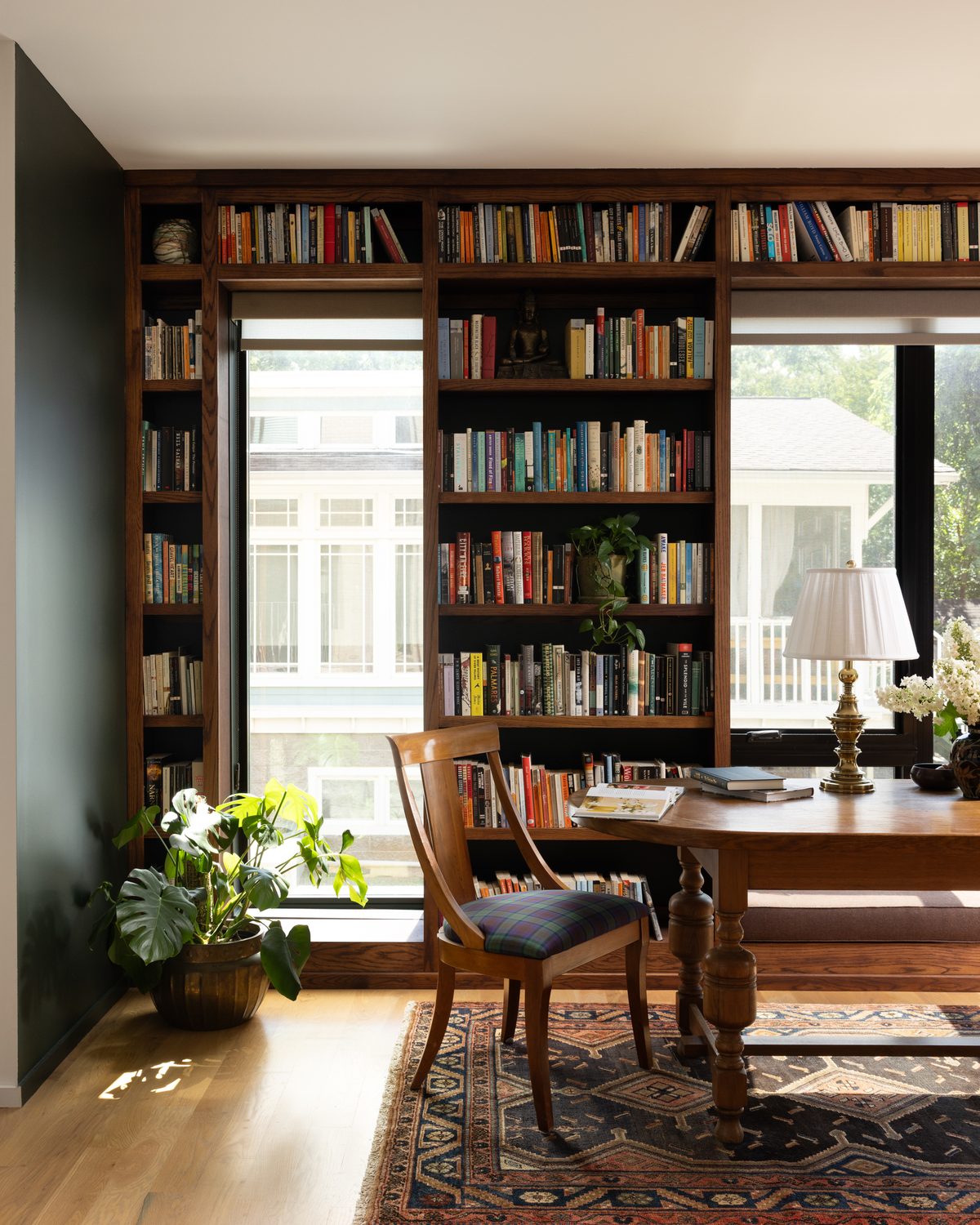 A library with floor-to-ceiling walnut bookshelves built around tall windows, a wood writing table with a brass lamp, and a plaid-upholstered chair