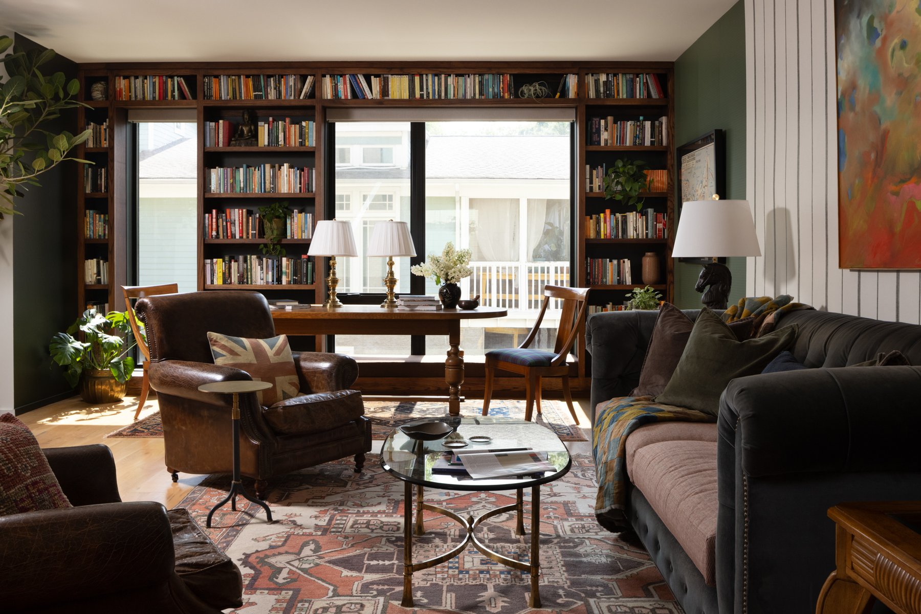 The library wall: walnut built-in bookshelves surrounding the windows, a library table with brass lamps, leather club chairs, and layered vintage rugs