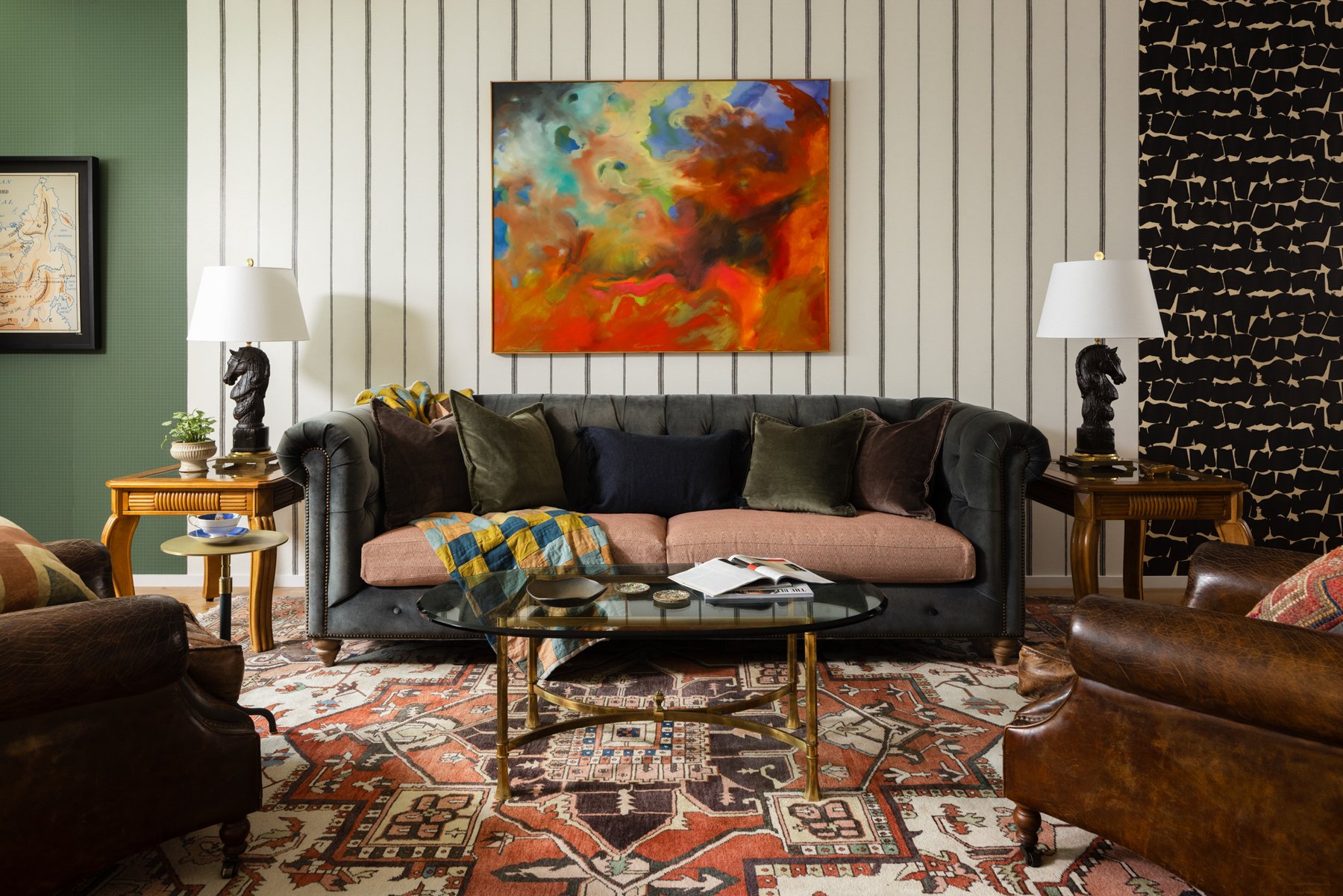A charcoal velvet chesterfield with a pink seat cushion under a large fiery abstract painting, flanked by striped and black-and-white wallpapered walls, horse-head lamps, and a vintage rug