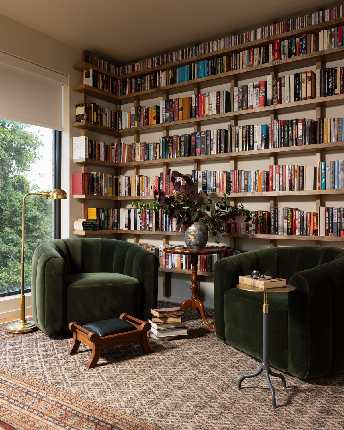 A reading corner in his office: two green velvet barrel chairs and a green leather footstool on a vintage rug, with a brass floor lamp and stacked books before a wall of open wood shelves full of books
