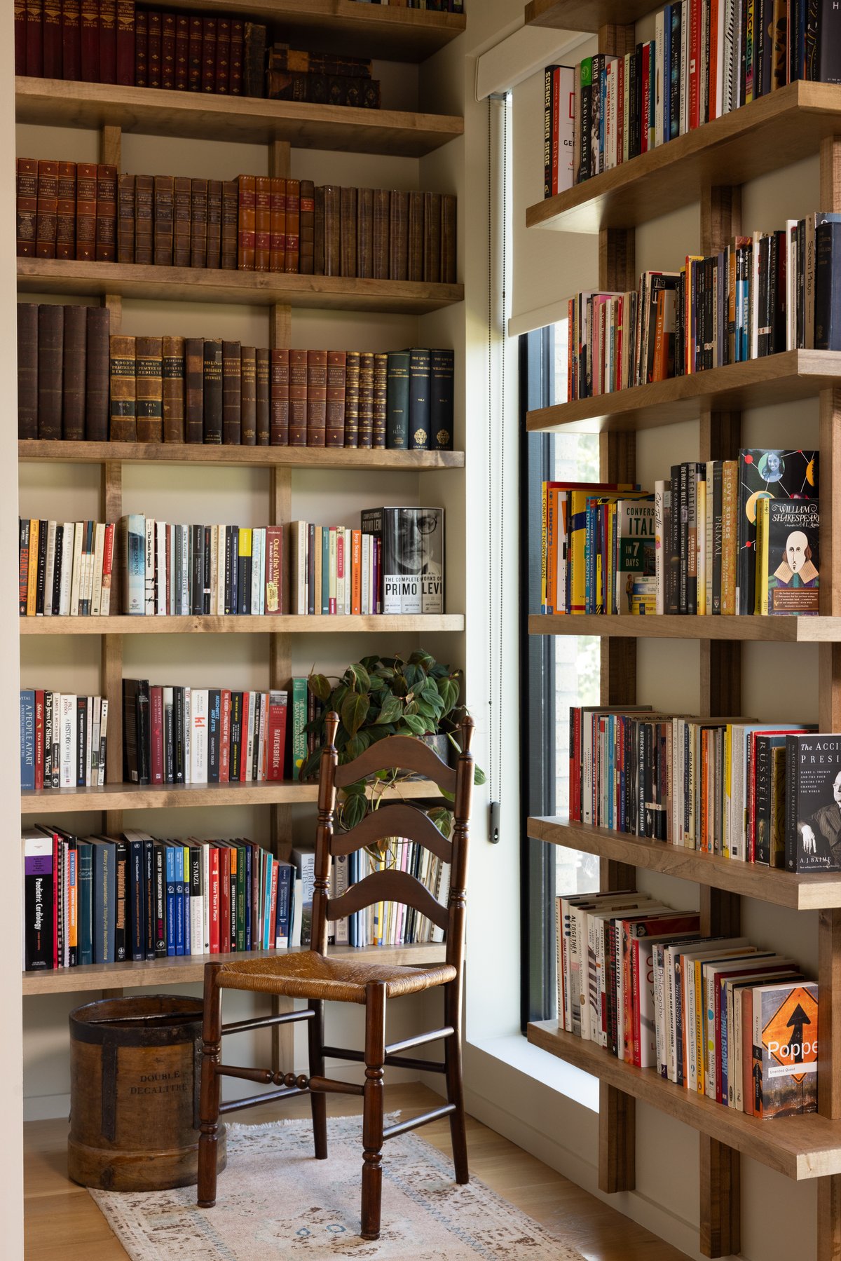 A corner of the office shelves: antique leather-bound volumes above rows of modern paperbacks, with a rush-seat ladder-back chair and a vintage wooden measure