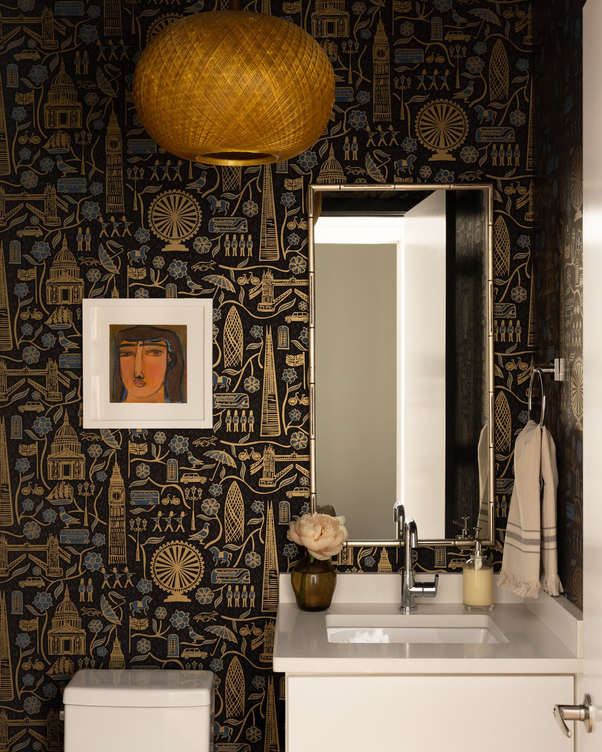The powder room wrapped in black and gold London-motif wallpaper with a woven pendant light, a bamboo-framed mirror, and a small framed portrait