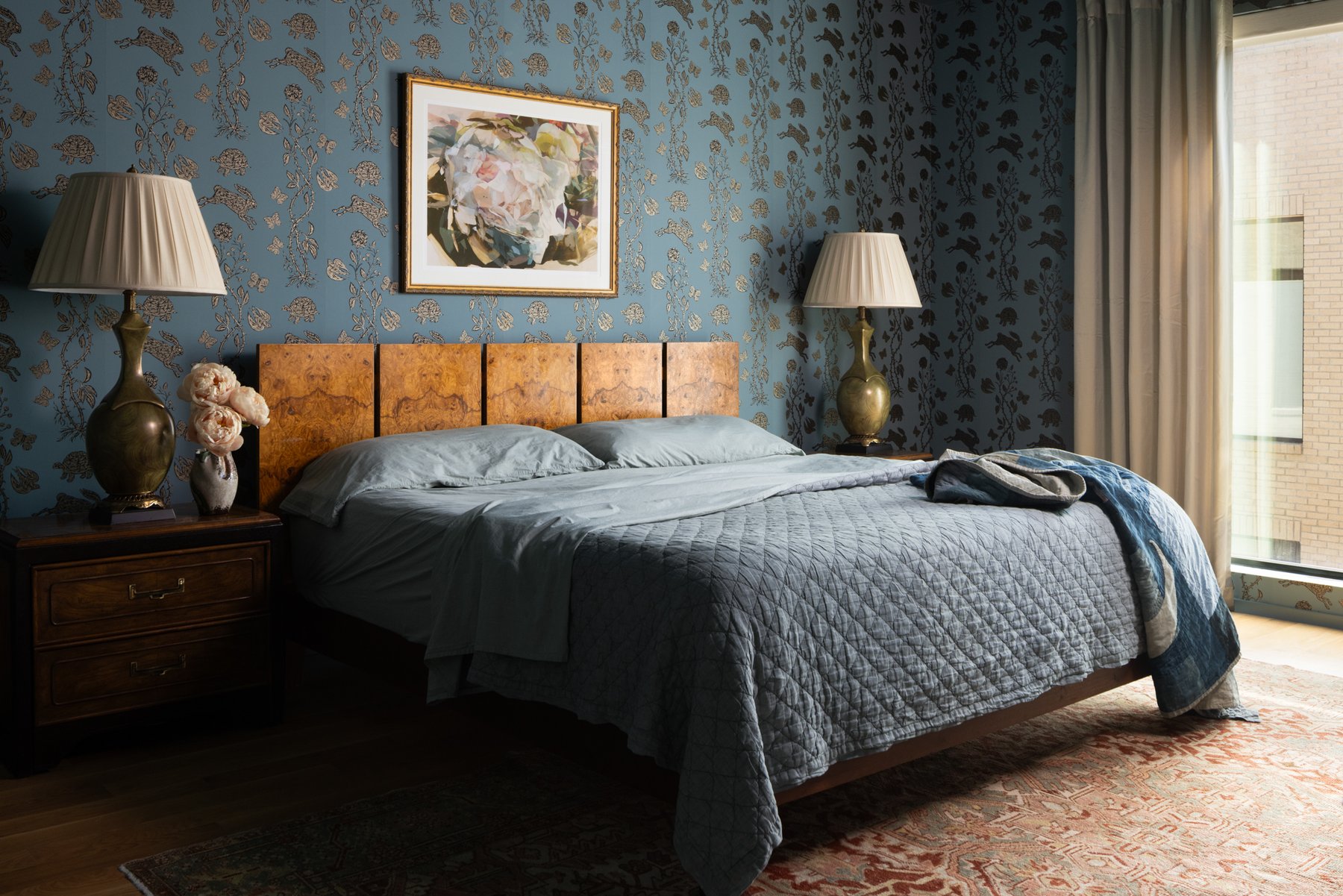 The primary bedroom: slate-blue hare-and-tortoise wallpaper, a burl wood headboard, vintage nightstands with sculptural green lamps, and a soft blue quilted bed
