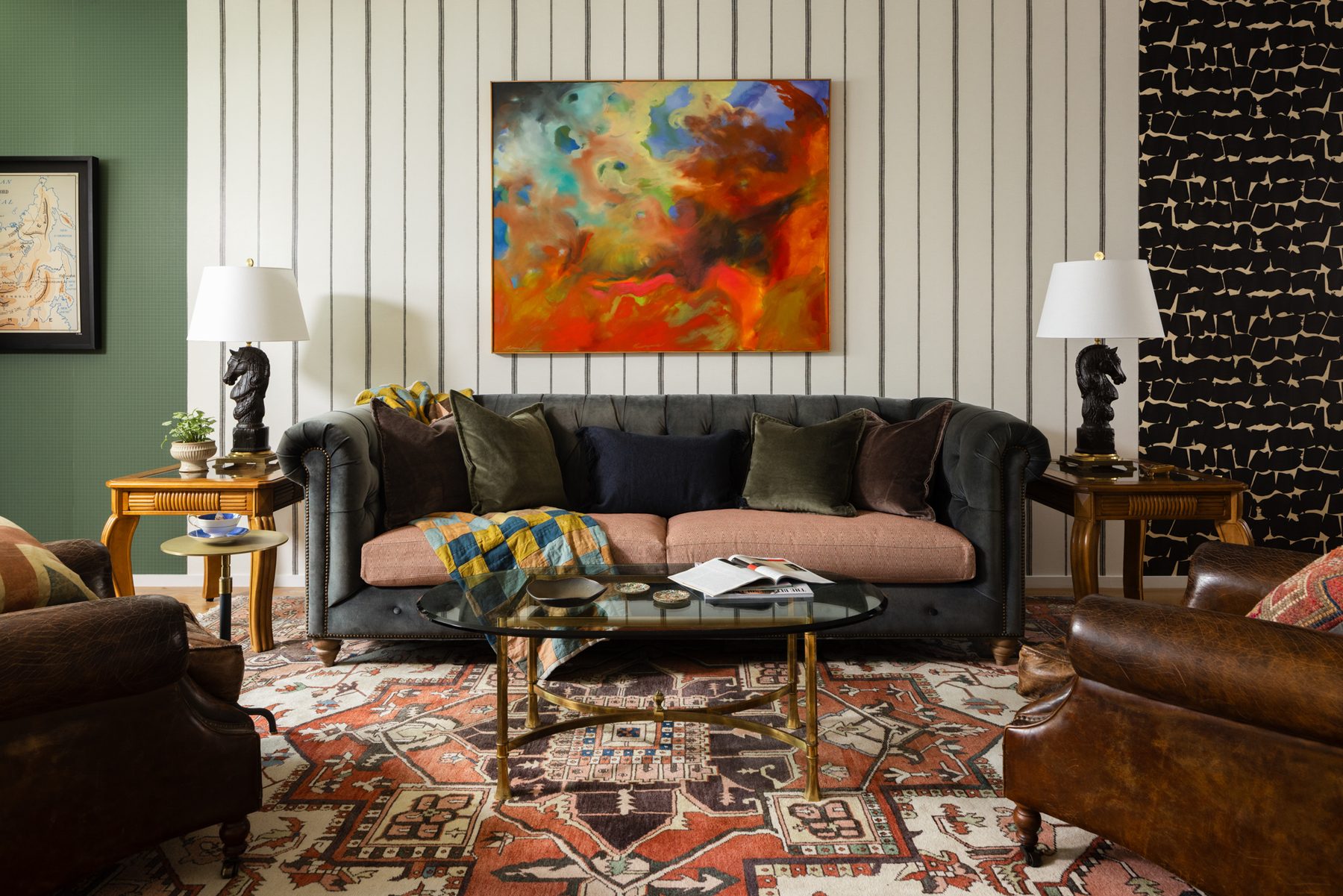 A charcoal velvet chesterfield sofa with a custom blush cushion, flanked by black horse lamps on vintage side tables, beneath a large abstract painting on striped wallpaper, with leather club chairs and a vintage rug