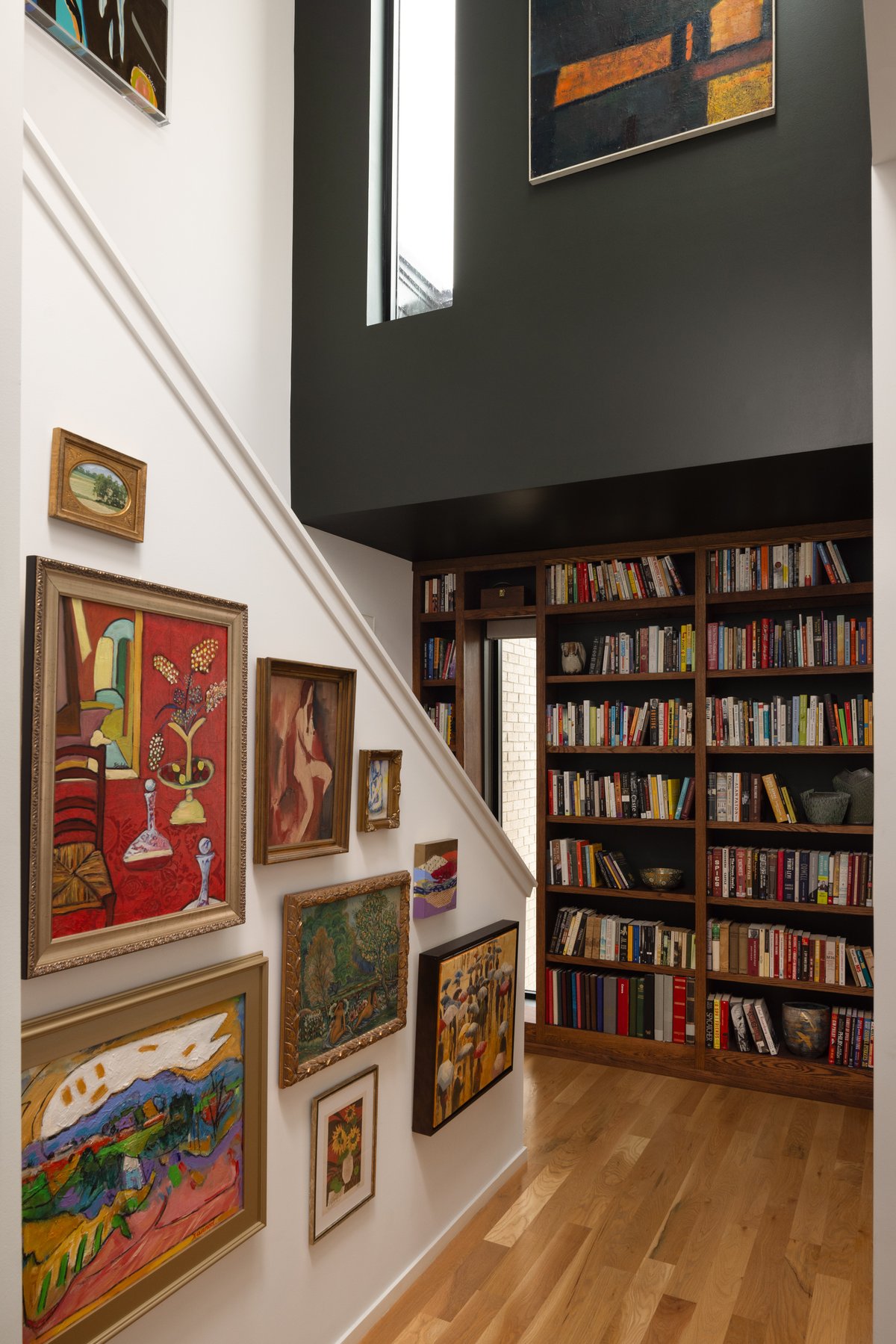 The stairwell as a gallery: a wall of collected paintings in gold frames rising alongside white stairs, with built-in bookshelves and a dark accent wall above