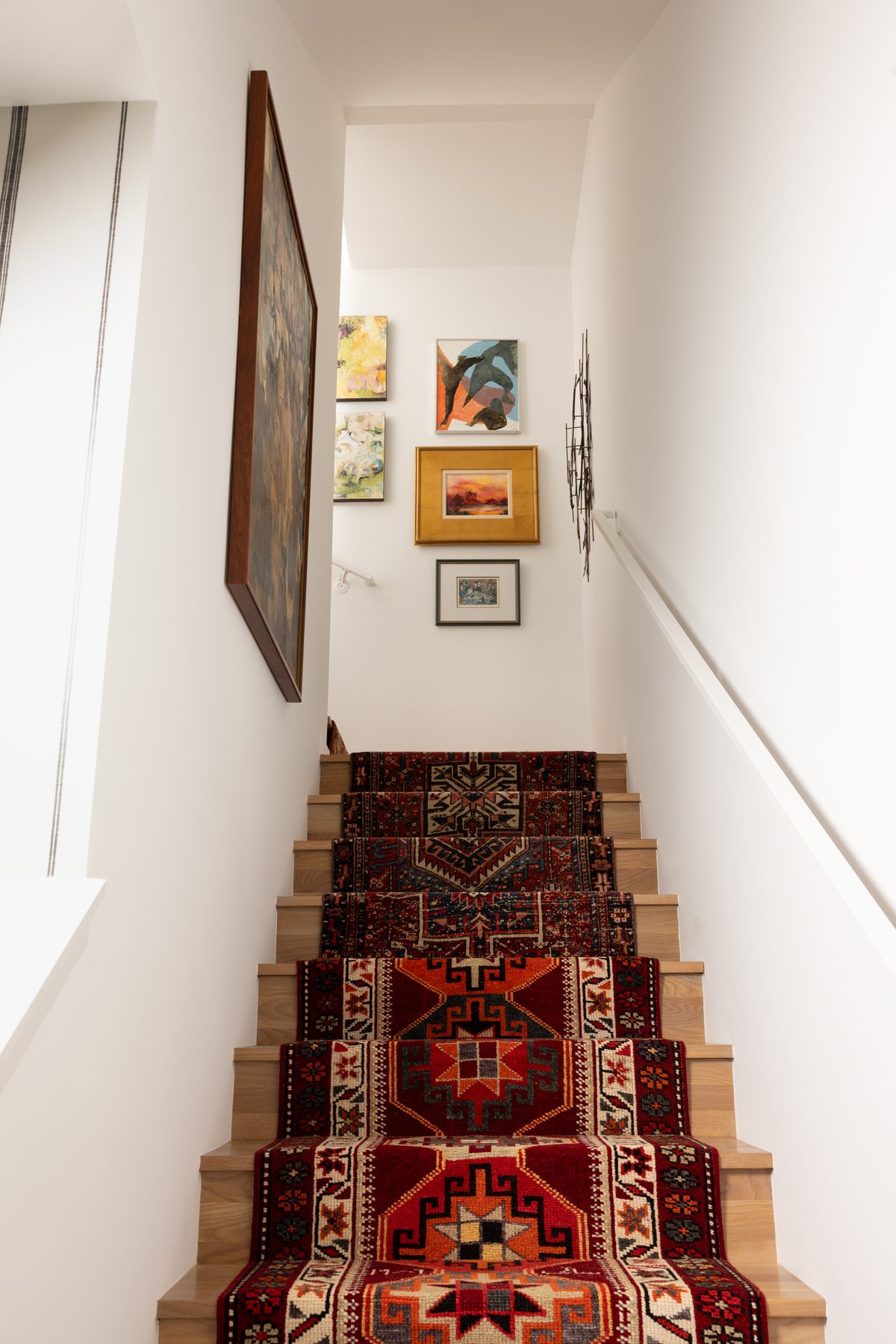 A staircase climbing between white walls hung with collected art, its steps covered in a patchwork runner of vintage rugs in deep reds