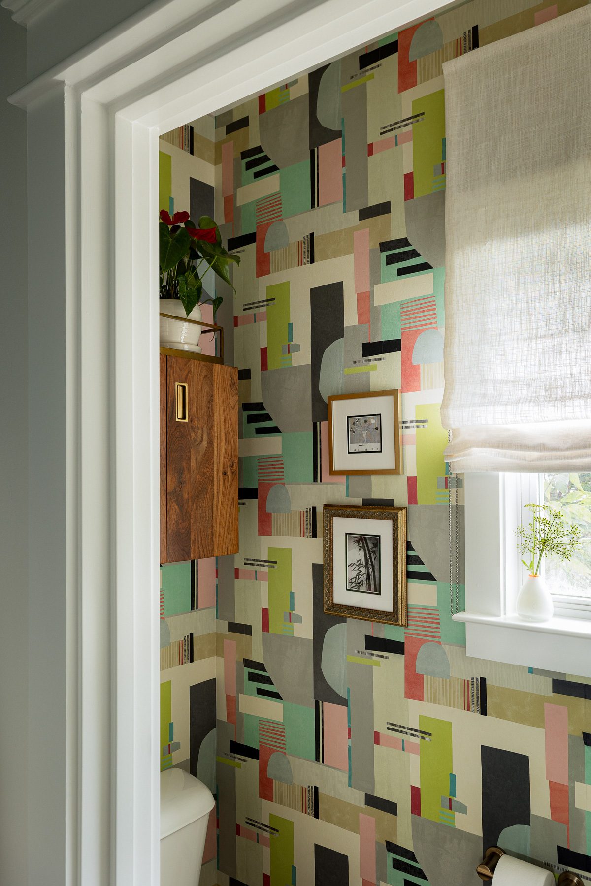 A water closet with colorful geometric wallpaper, a walnut wall cabinet, framed art, and a linen roman shade