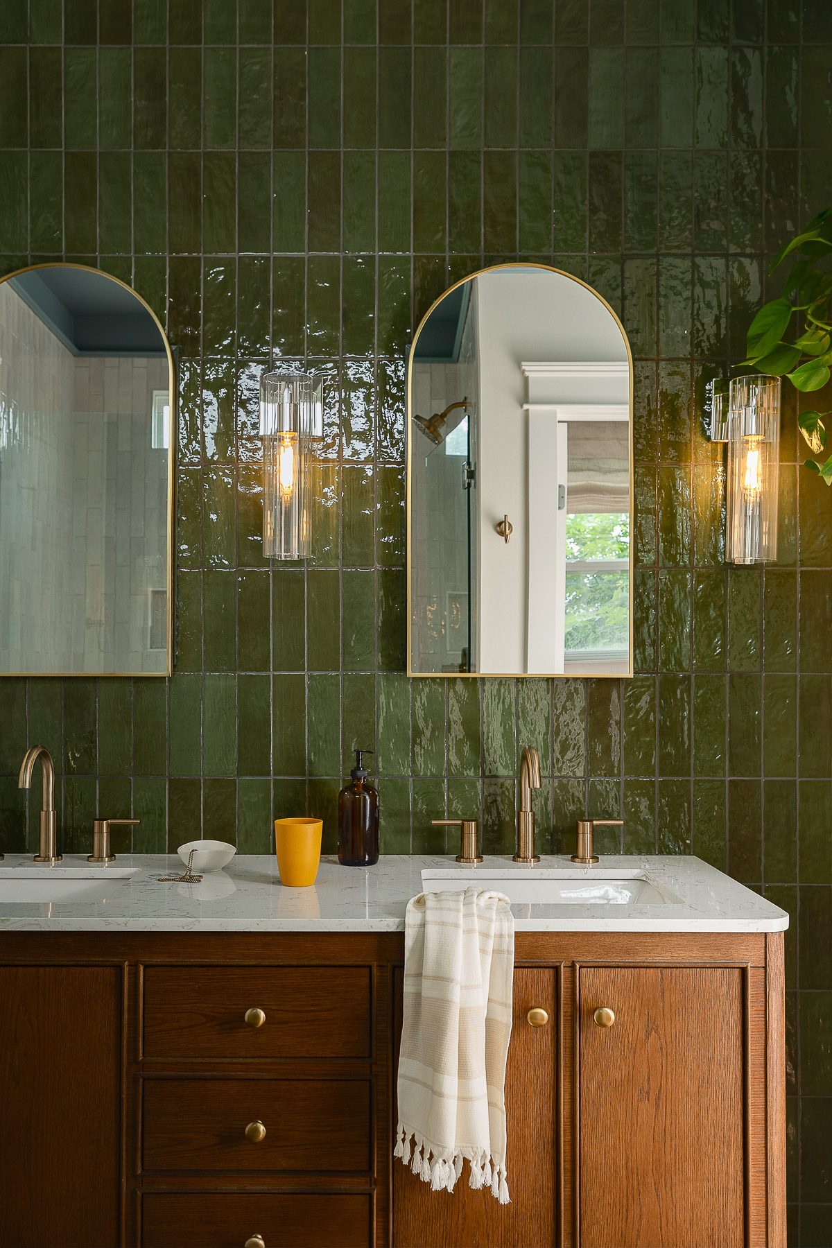 A double vanity with green zellige tile, arched brass mirrors, tubular sconces, and warm oak cabinetry