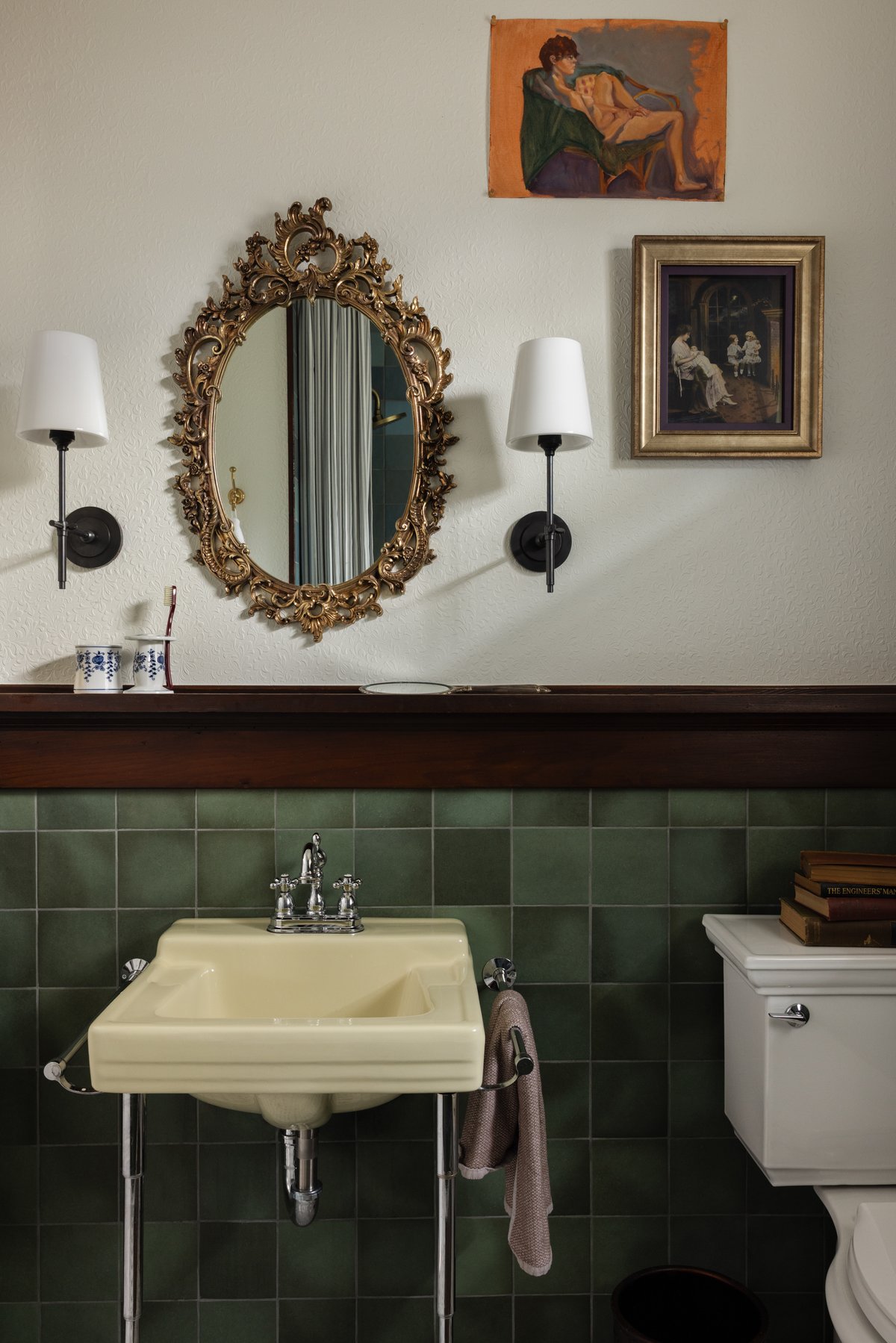 The vanity wall: an ornate gilt mirror between black sconces over the butter-yellow sink, with a figure study pinned to the wall and green tile below