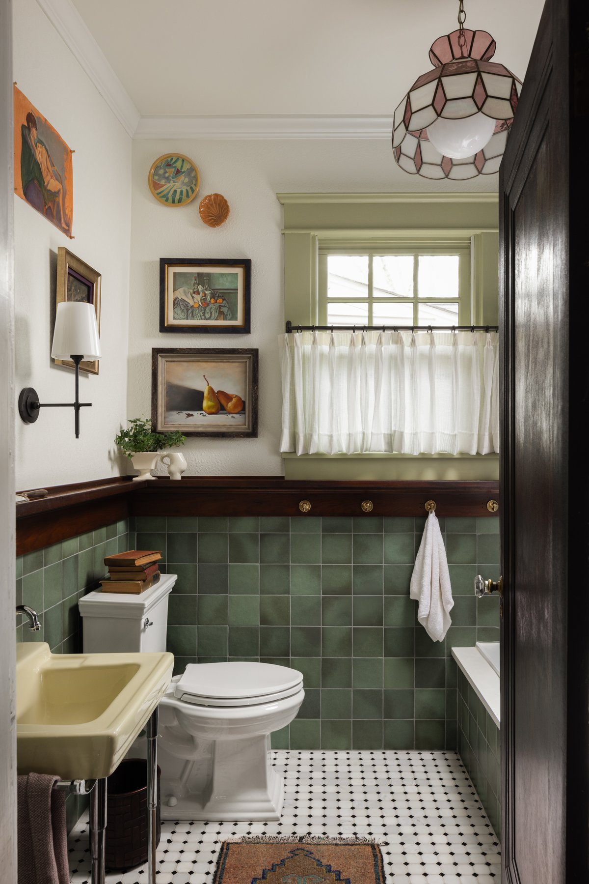 The bathroom: green square tile under a dark wood ledge with brass hooks, a butter-yellow vintage sink, black-and-white mosaic floor, cafe curtains on a green window, and a stained-glass pendant