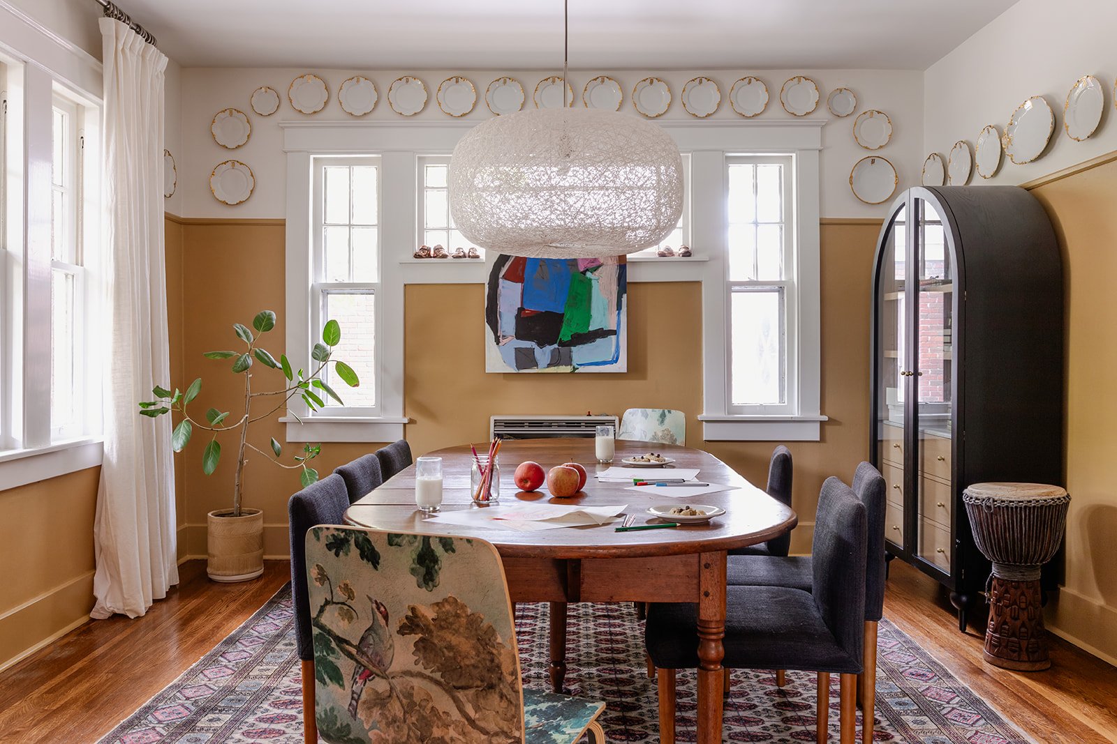 The dining room ringed with gold-rimmed plates high on the wall, an antique oval table with mismatched chairs including a bird-print bentwood, a woven pendant, a black arched cabinet, and a djembe drum in the corner