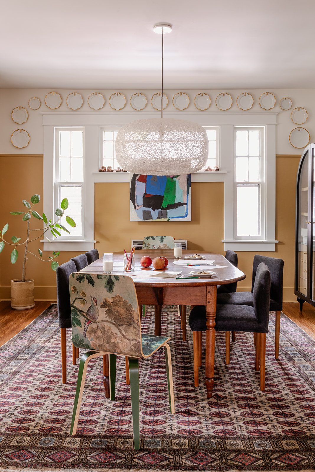 A dining room ringed with gold-rimmed plates on the wall, an antique oval table with mismatched chairs including botanical-print bentwood seats, and a woven pendant light