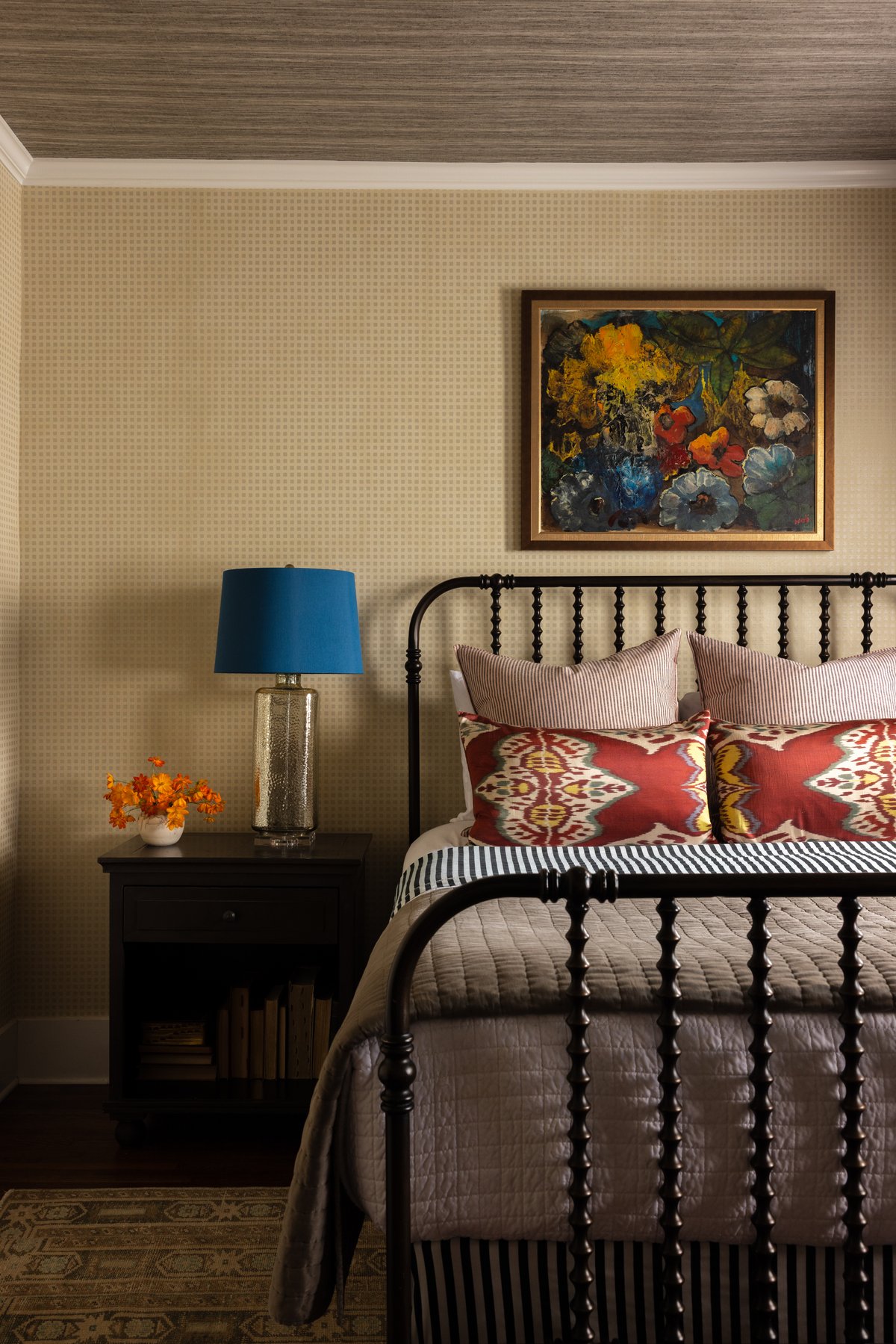 The guest bedroom: a black spool bed with striped and red ikat pillows under a moody floral painting, gingham wallpaper, a grasscloth ceiling, and a blue-shaded mercury glass lamp