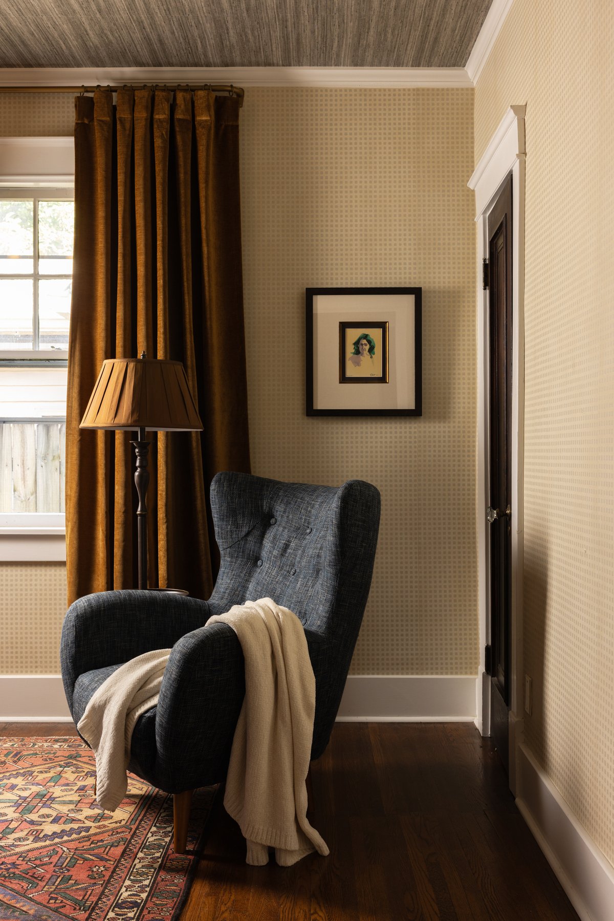 A corner of the guest bedroom: a navy tufted wingback with a cream throw beside amber velvet drapes, a pleated-shade floor lamp, and a small green portrait in a double frame