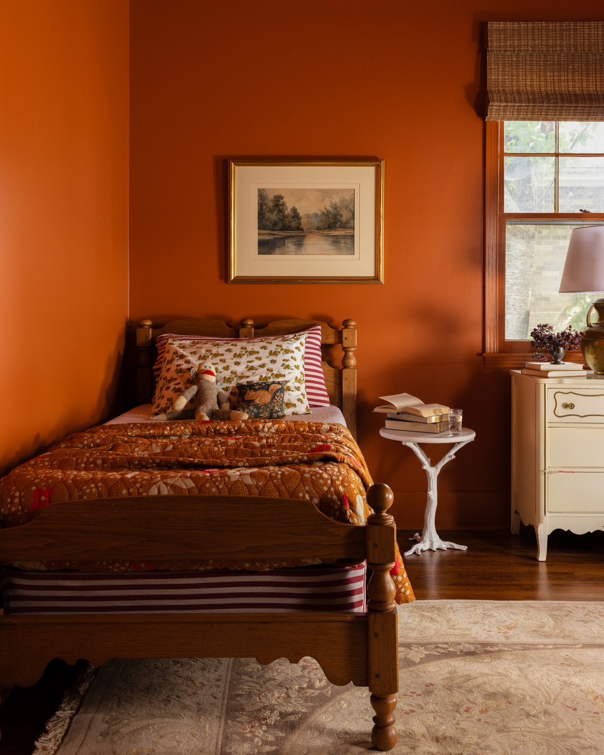The third bed: a turned-wood twin with a rust floral quilt and sock monkey, a faux-bois side table stacked with books, and a gold-framed landscape on the orange wall