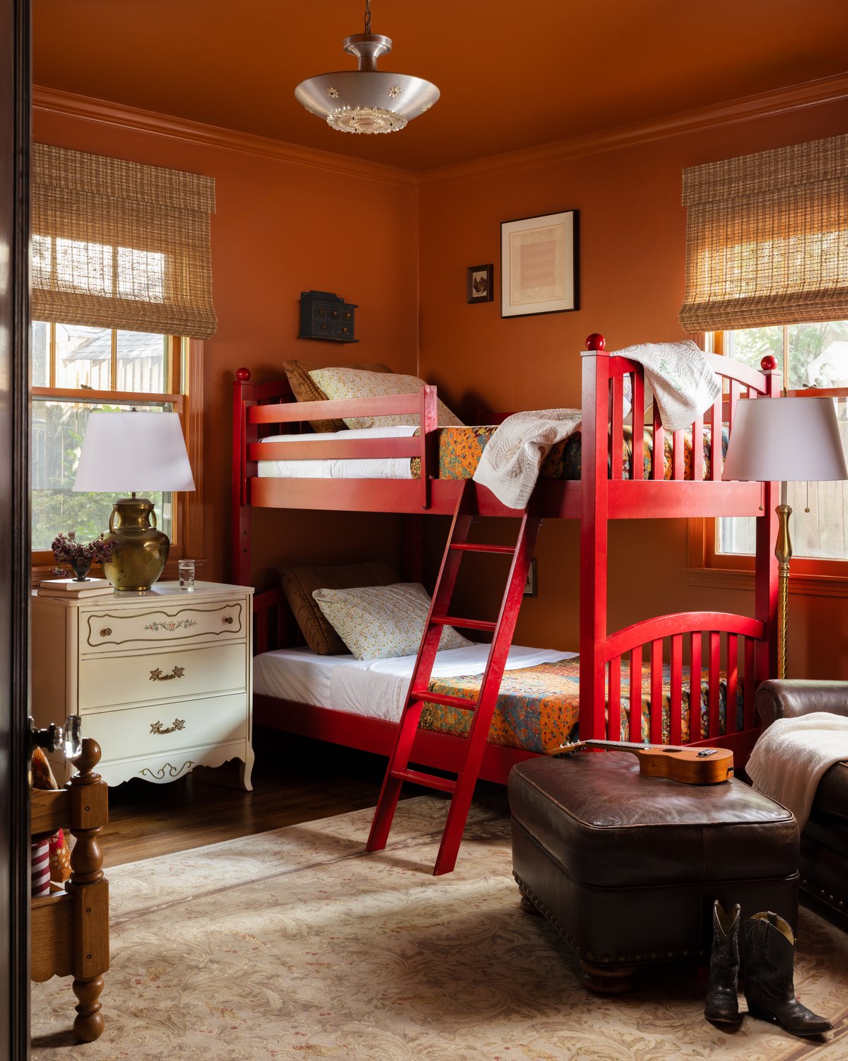 The kids&rsquo; room today: cherry-red bunk beds against burnt-orange walls and ceiling, a painted vintage dresser with a brass lamp, bamboo shades, and a leather ottoman with a ukulele