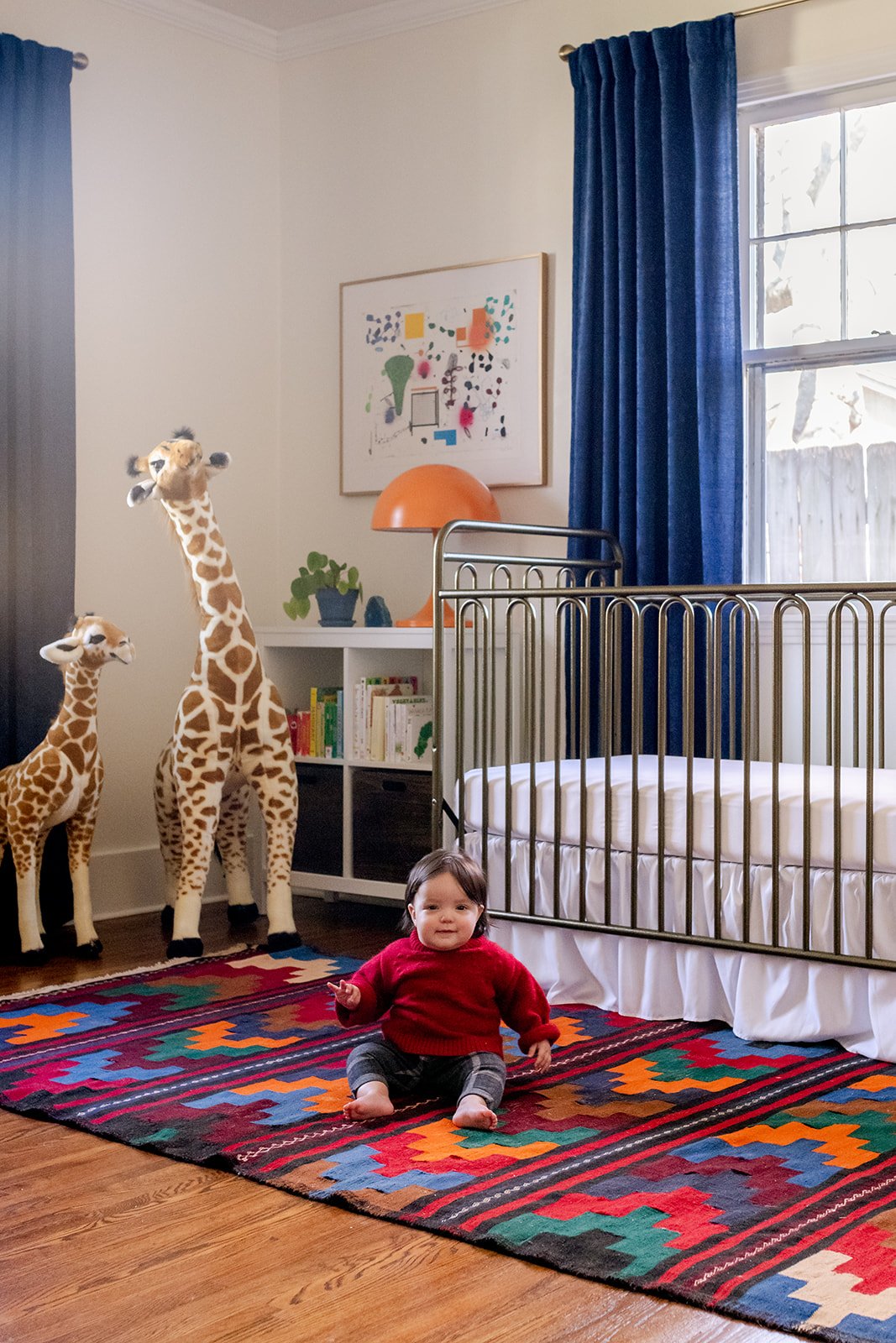 The 2021 nursery: a brass crib by navy velvet drapes, a colorful vintage kilim with the baby sitting on it, plush giraffes, and an orange mushroom lamp on a white bookcase