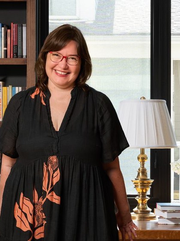 Kristin standing in a home library between walnut bookshelves and a tall window, a brass lamp beside her